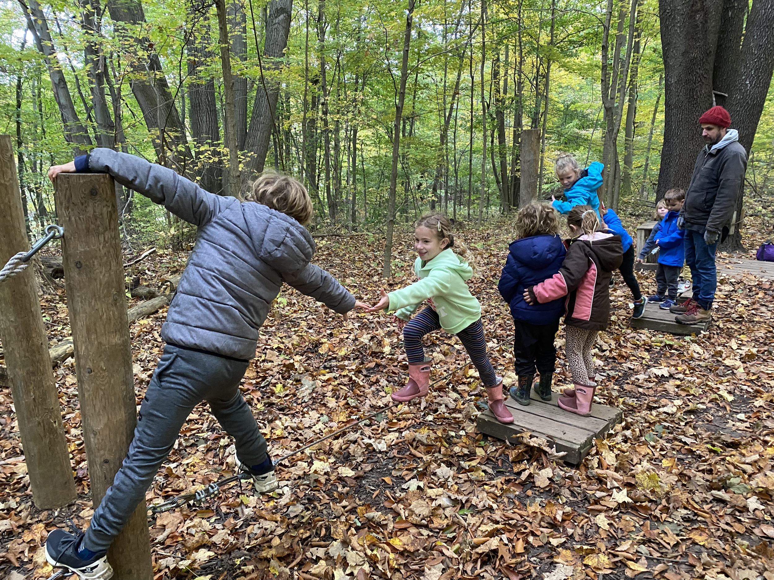 Children and a man playing on a make-believe bridge in a forest during fall, with leaves on the ground and trees with changing leaves.