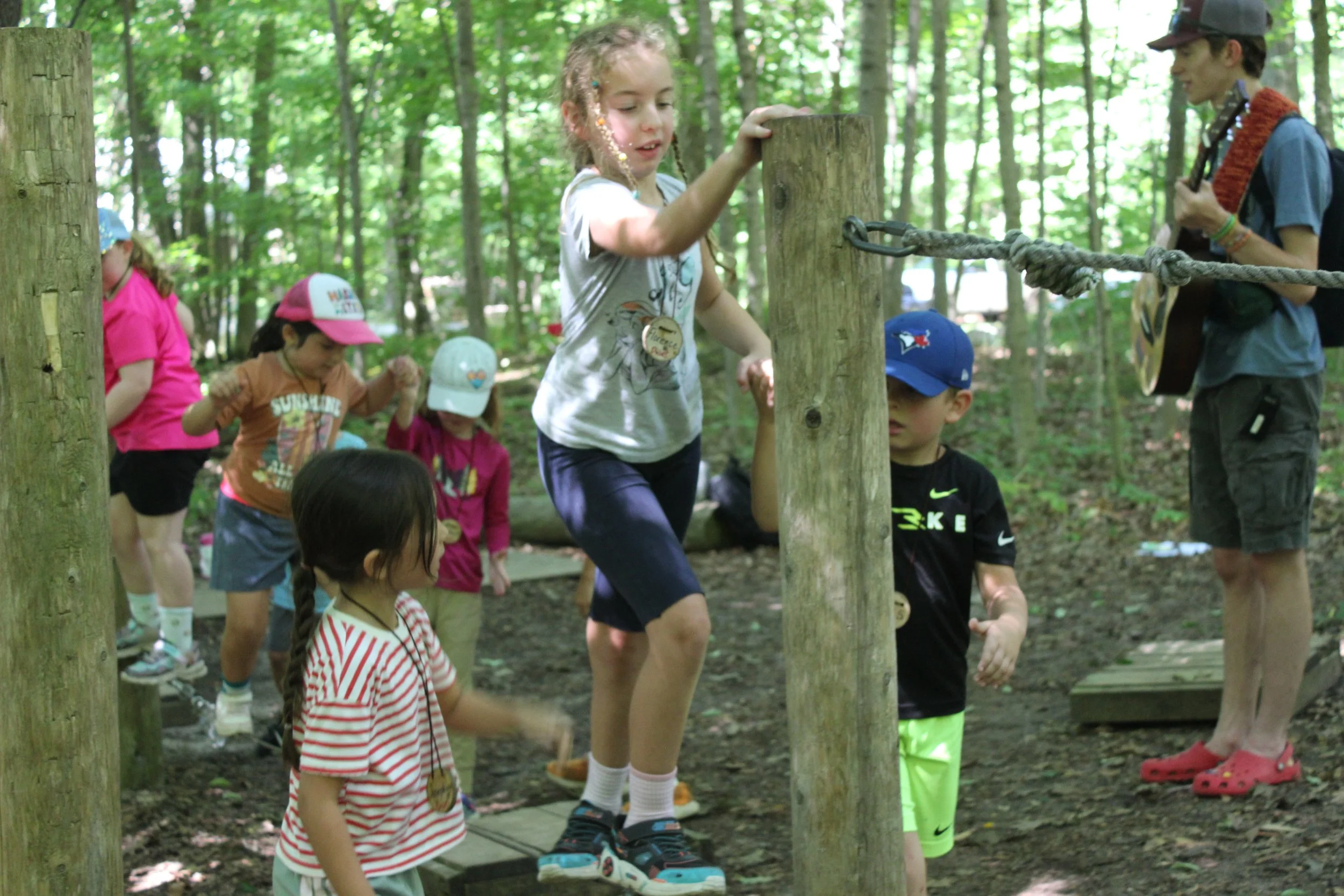 Children playing on an obstacle course in a wooded forest, with an adult supervising and playing guitar.