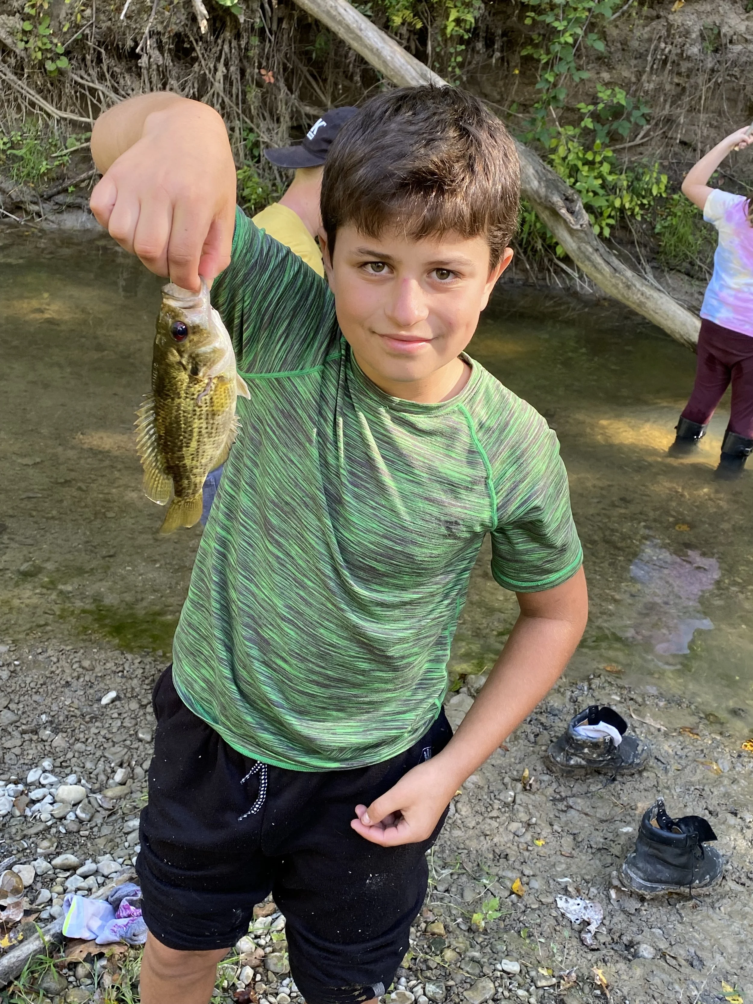 A young boy holding a small fish with a happy expression, standing near a creek in a green and black shirt, with other children in the background.