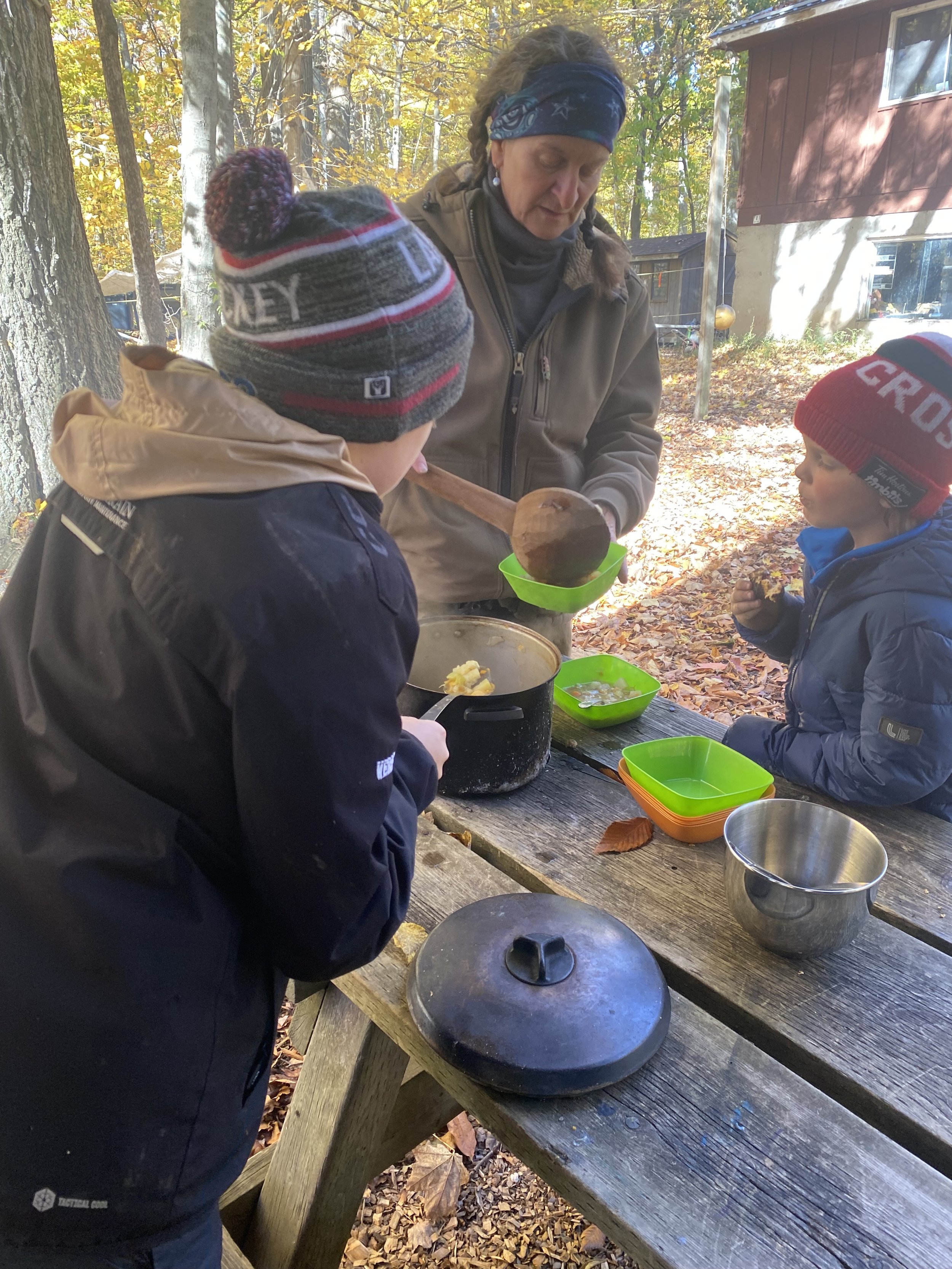 An older woman and two young boys with winter hats and jackets are preparing food outdoors at a wooden picnic table in a wooded area during autumn.