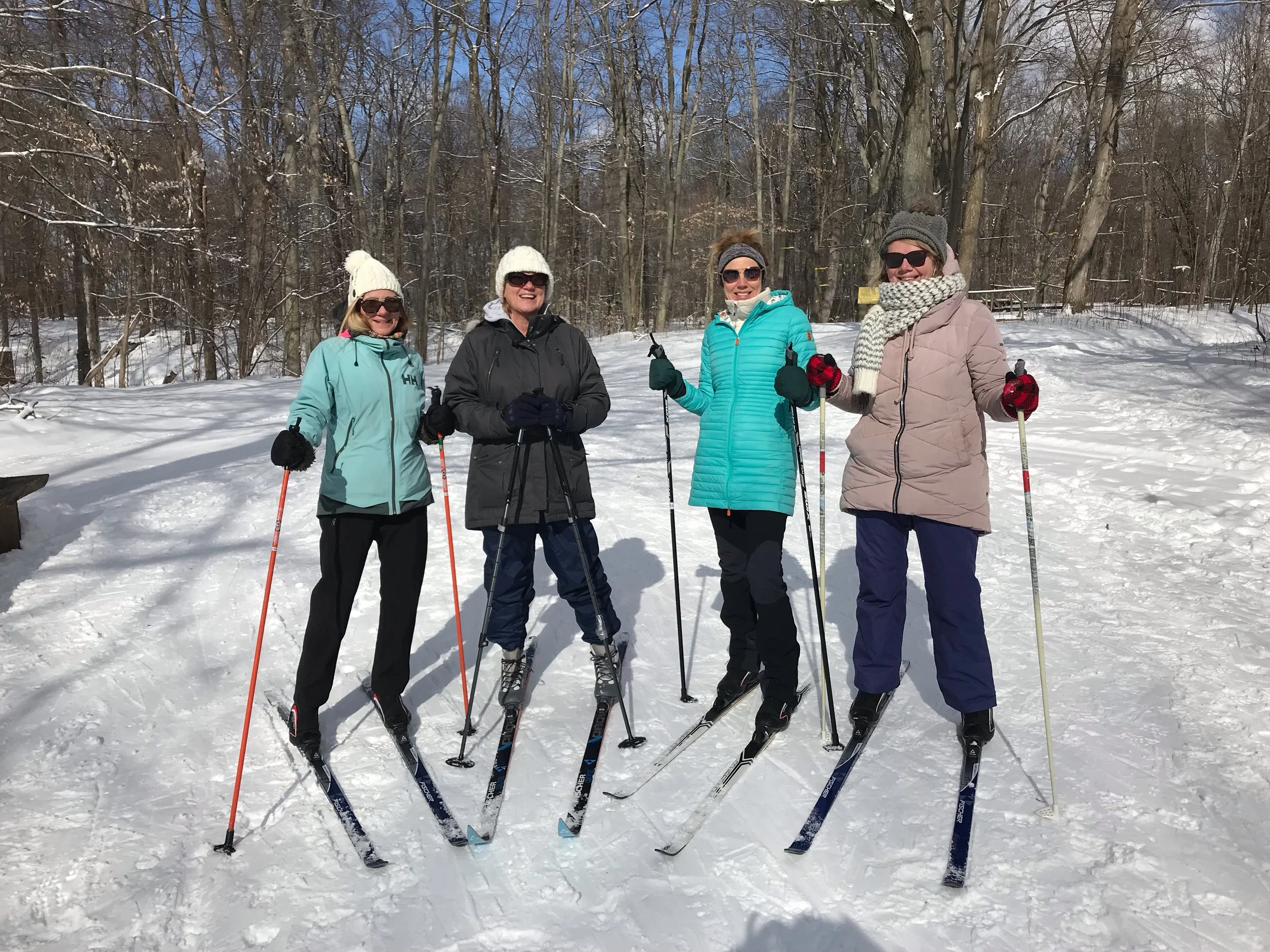 Four women standing on snow with ski poles and skis, dressed in winter jackets, hats, and sunglasses, in a snowy wooded area.