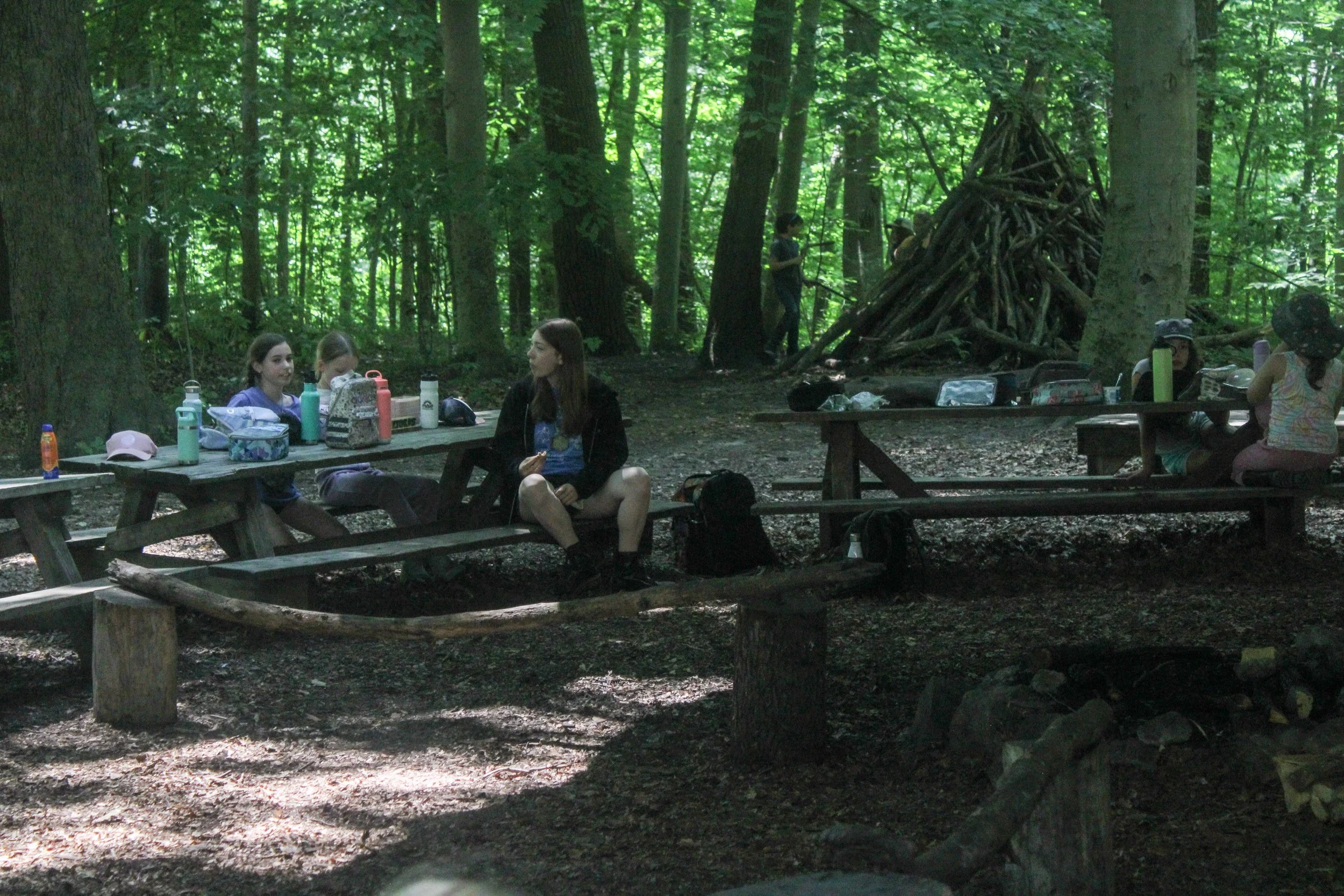 A group of children sitting at a wooden picnic table in a forested area with trees and a teepee made of branches in the background.