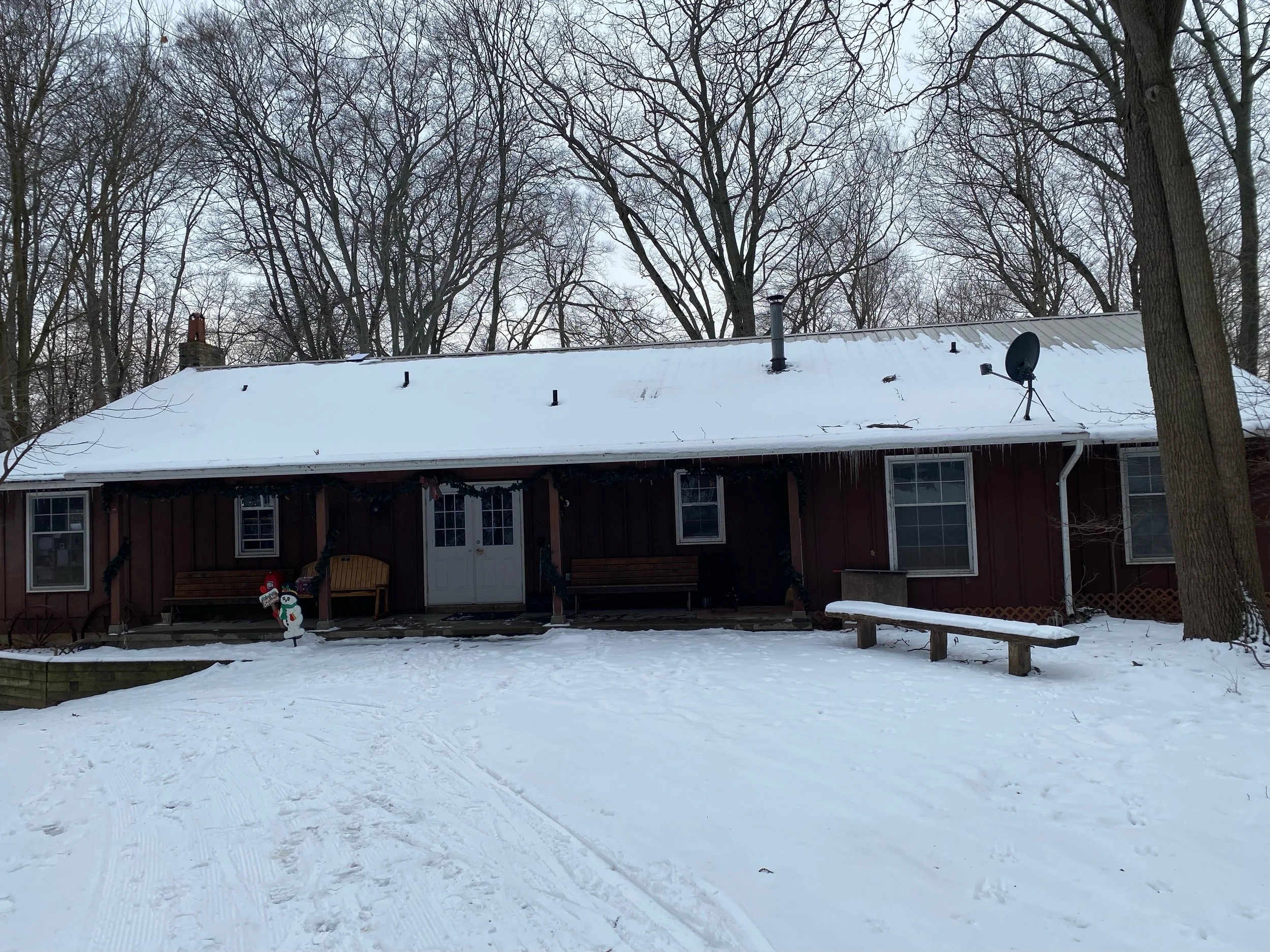 Red house in winter with snow-covered yard and leafless trees in the background.