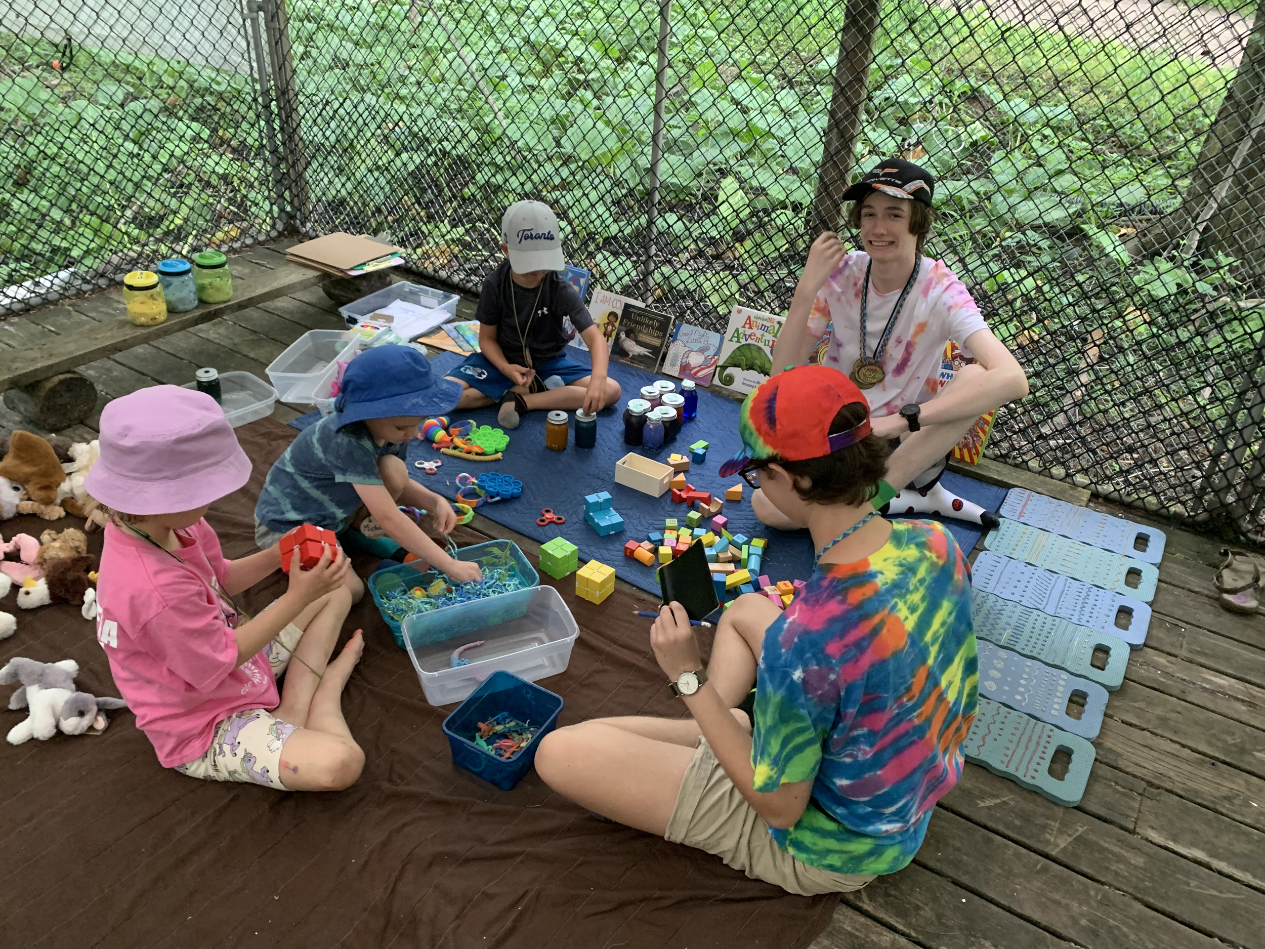 A group of children and teenagers sitting on a wooden deck, engaged in arts and crafts activities with various toys, puzzles, and art supplies. They are outdoors with a mesh fence and green foliage in the background.