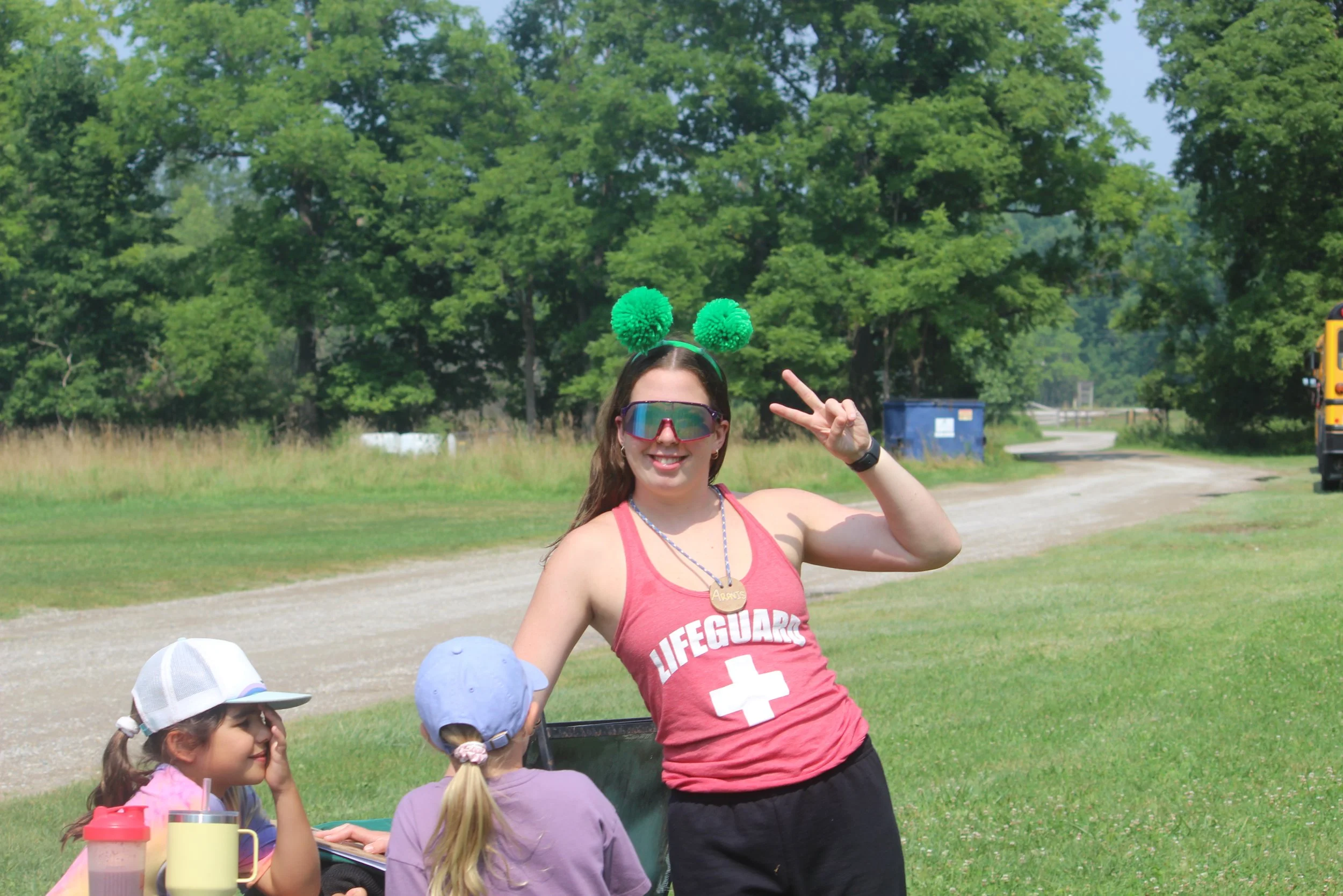 Lifeguard wearing a red tank top with a white cross and green pom-pom headband making a peace sign near children sitting at a table outdoors with trees and a yellow bus in the background.