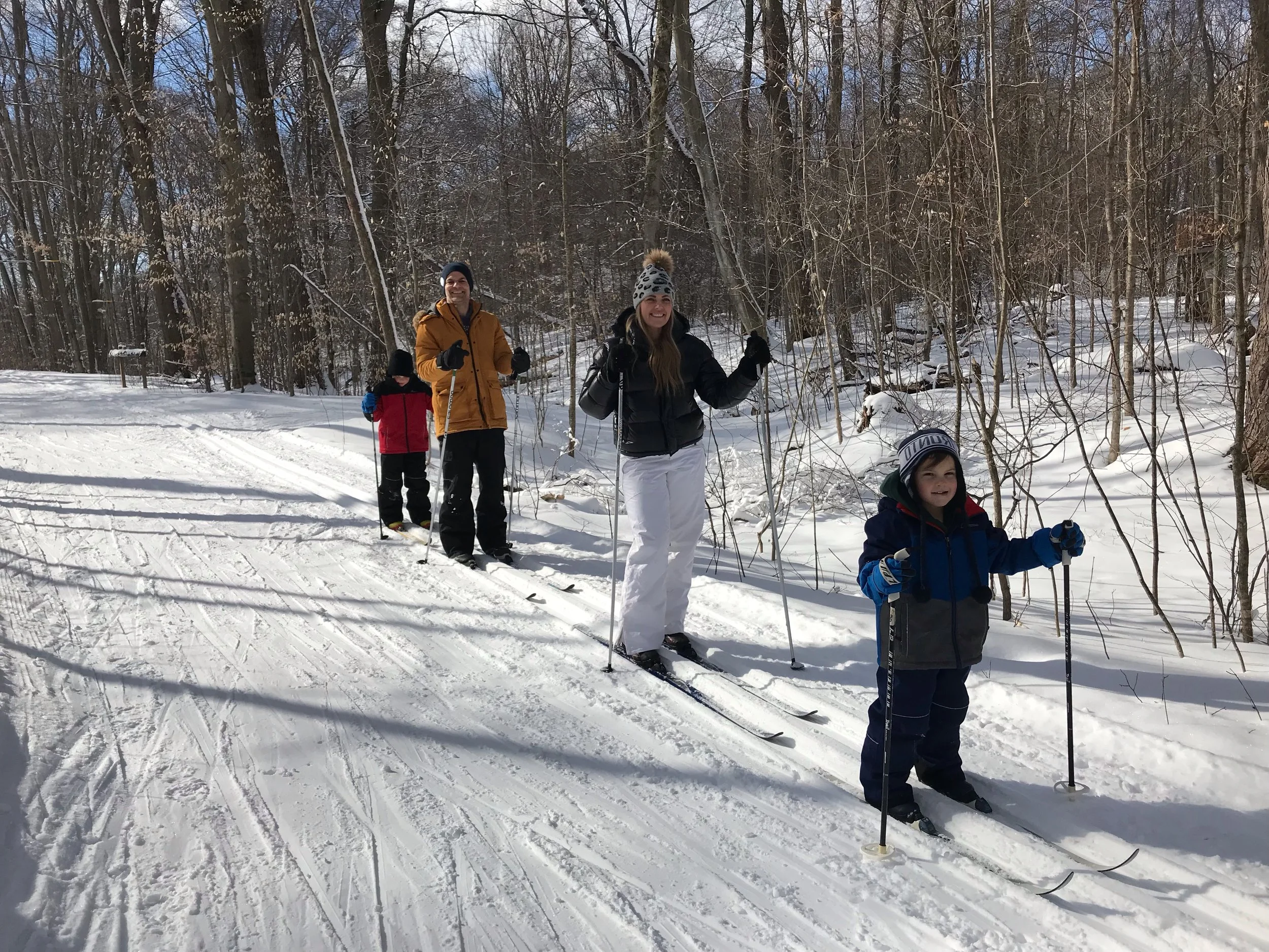 A family of four cross-country skiing on a snowy trail in a wooded area during winter, with leafless trees and snow-covered ground.