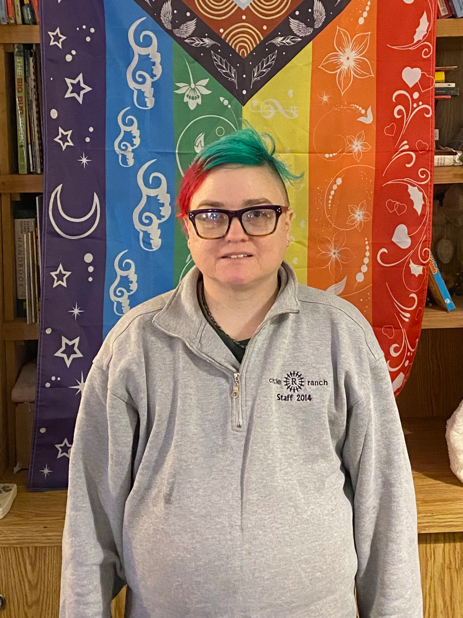 Person with short hair dyed in rainbow colors with green, blue, and red shades, wearing glasses, standing in front of a rainbow Pride flag with various designs, inside a room with bookshelves on the sides.