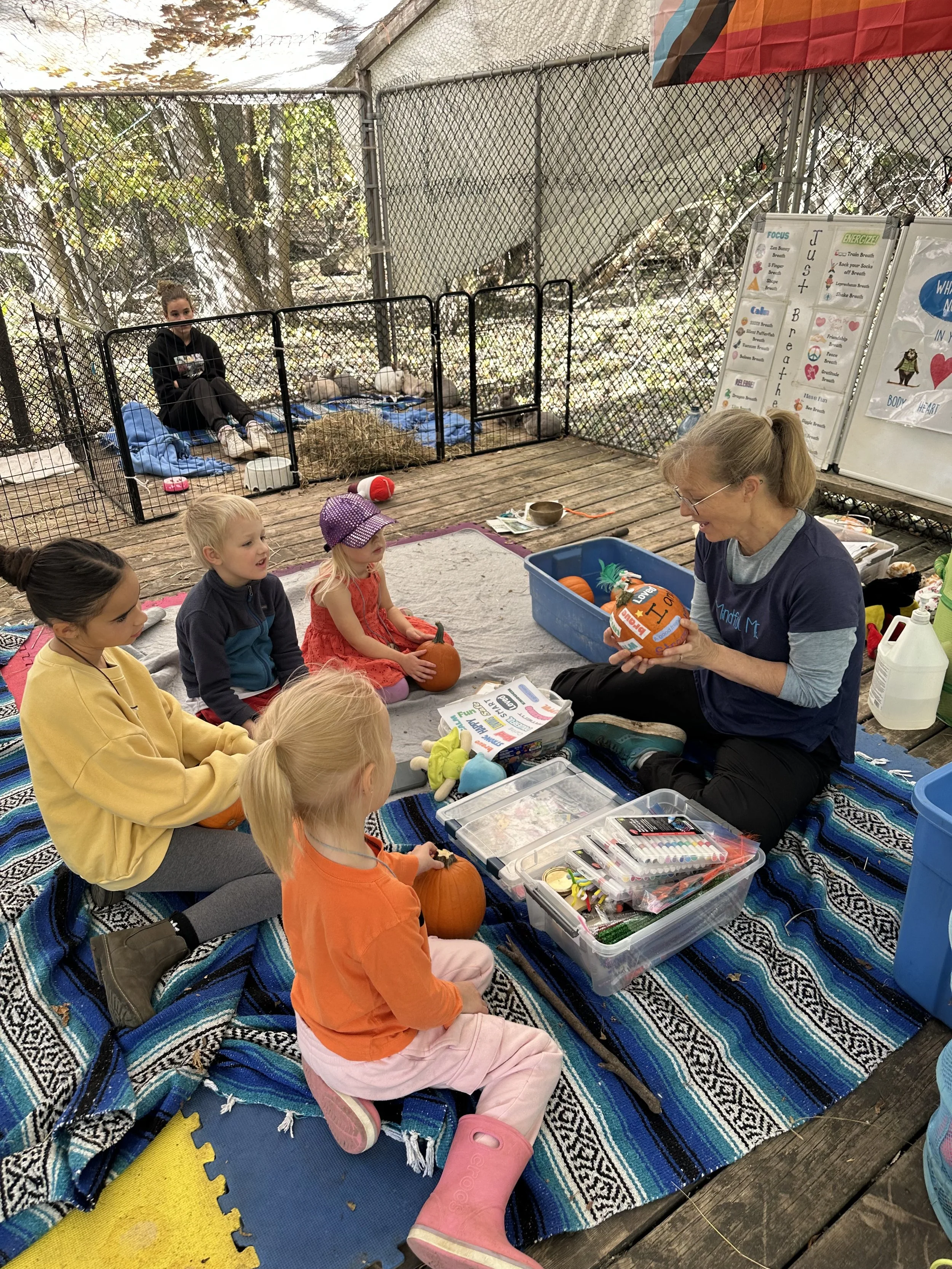 A woman teaching children about pumpkins in an outdoor setting with a fenced animal area in the background.