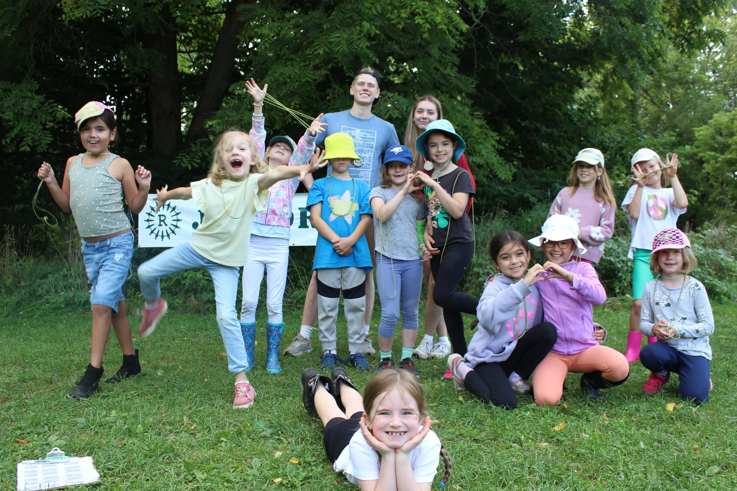 A group of children and two adults outdoors in a grassy area with trees in the background, smiling and posing for a photo.