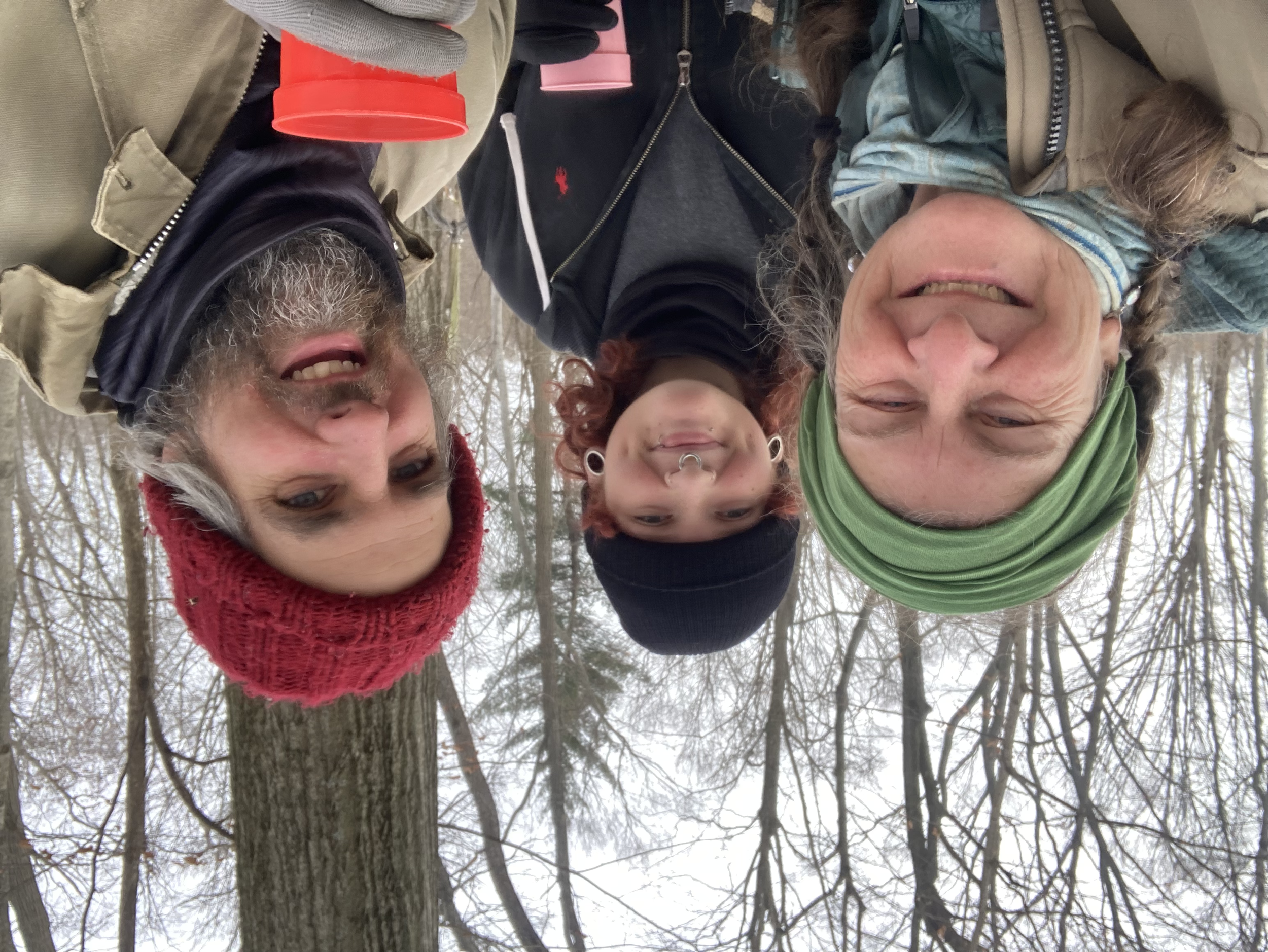 Four people smiling in a winter forest, wearing warm clothing and hats, with leafless trees and overcast sky in the background.