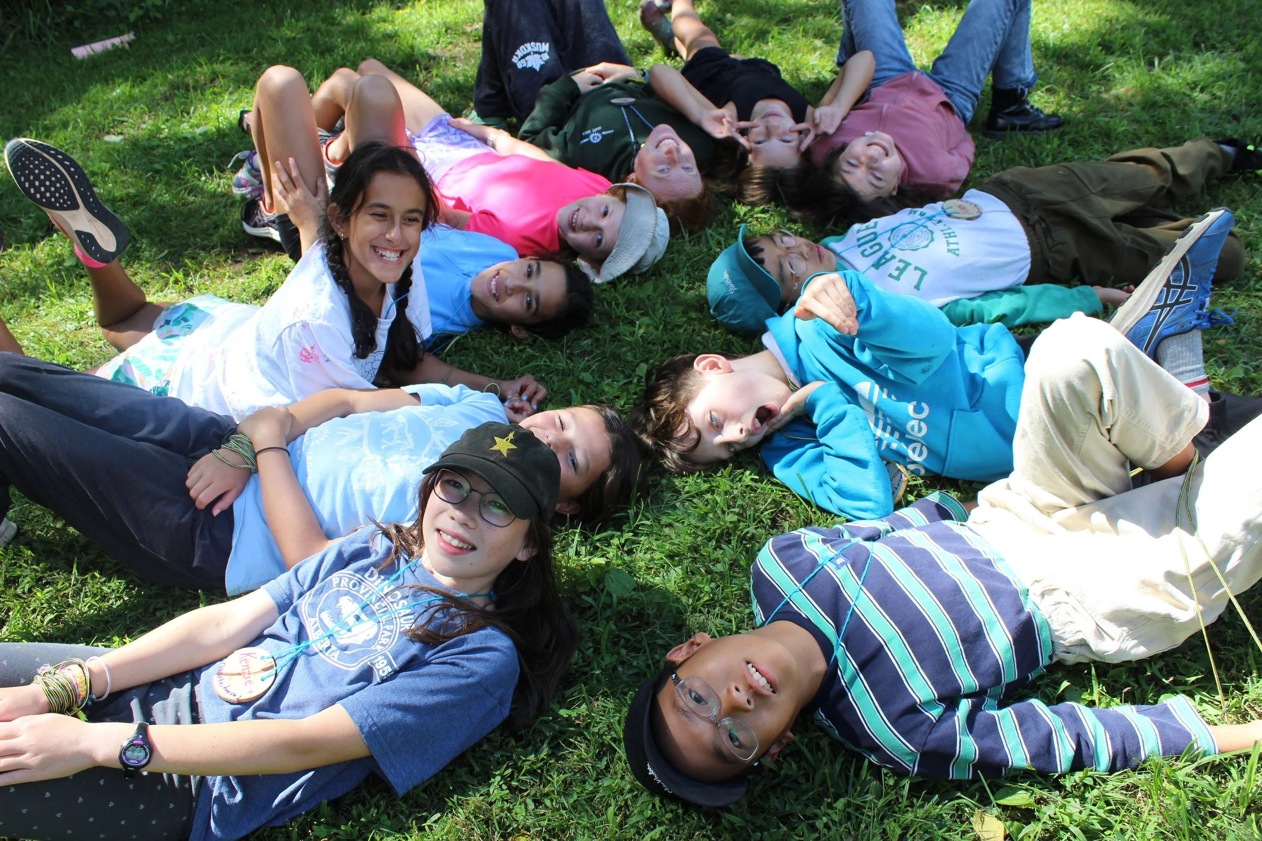 A group of children and teenagers lying on the grass in a circle outside, smiling and making silly faces.