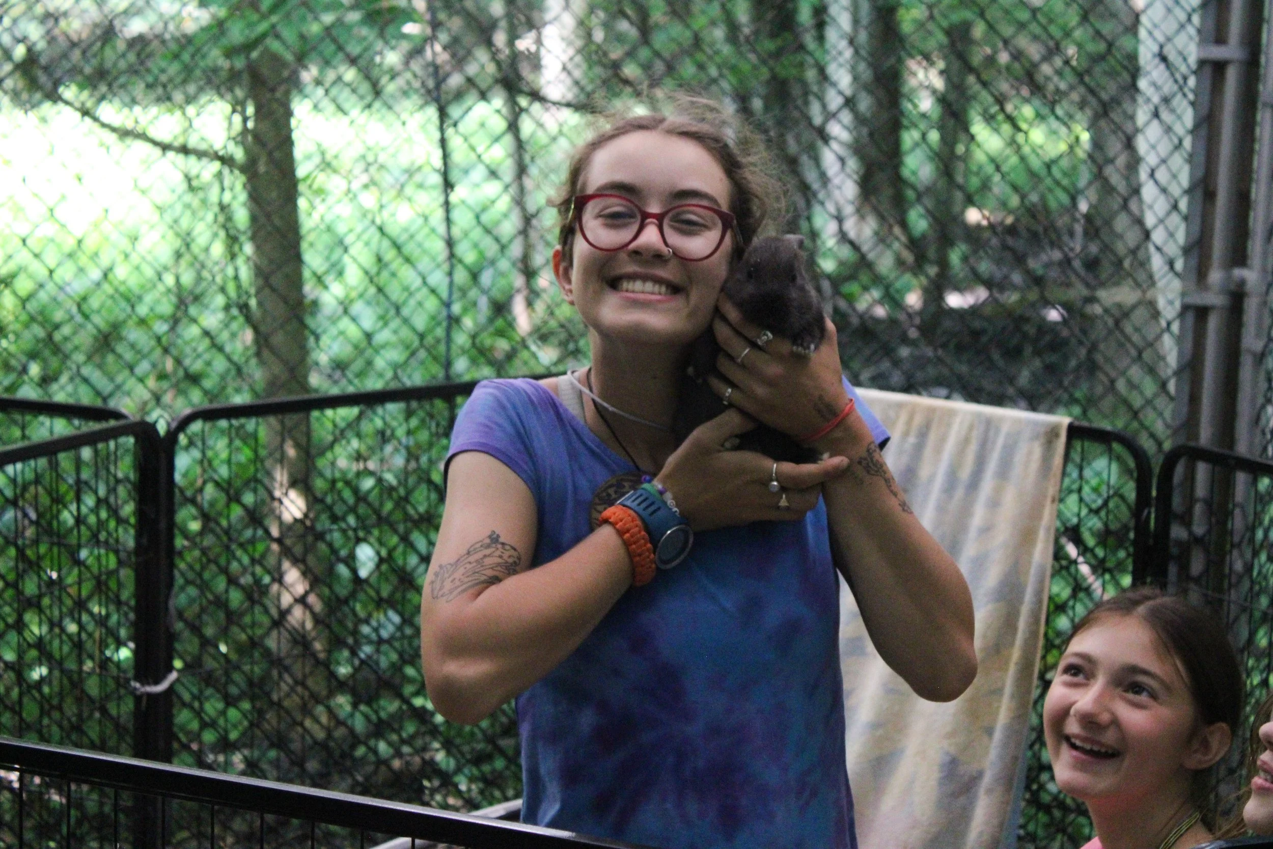 A smiling woman with glasses holding a small dark animal, likely a rabbit, in her hands. She is wearing a blue T-shirt and has tattoos on her arms. There are two young girls smiling in the foreground, one of whom is looking up at her. The background shows a fenced outdoor area with greenery.