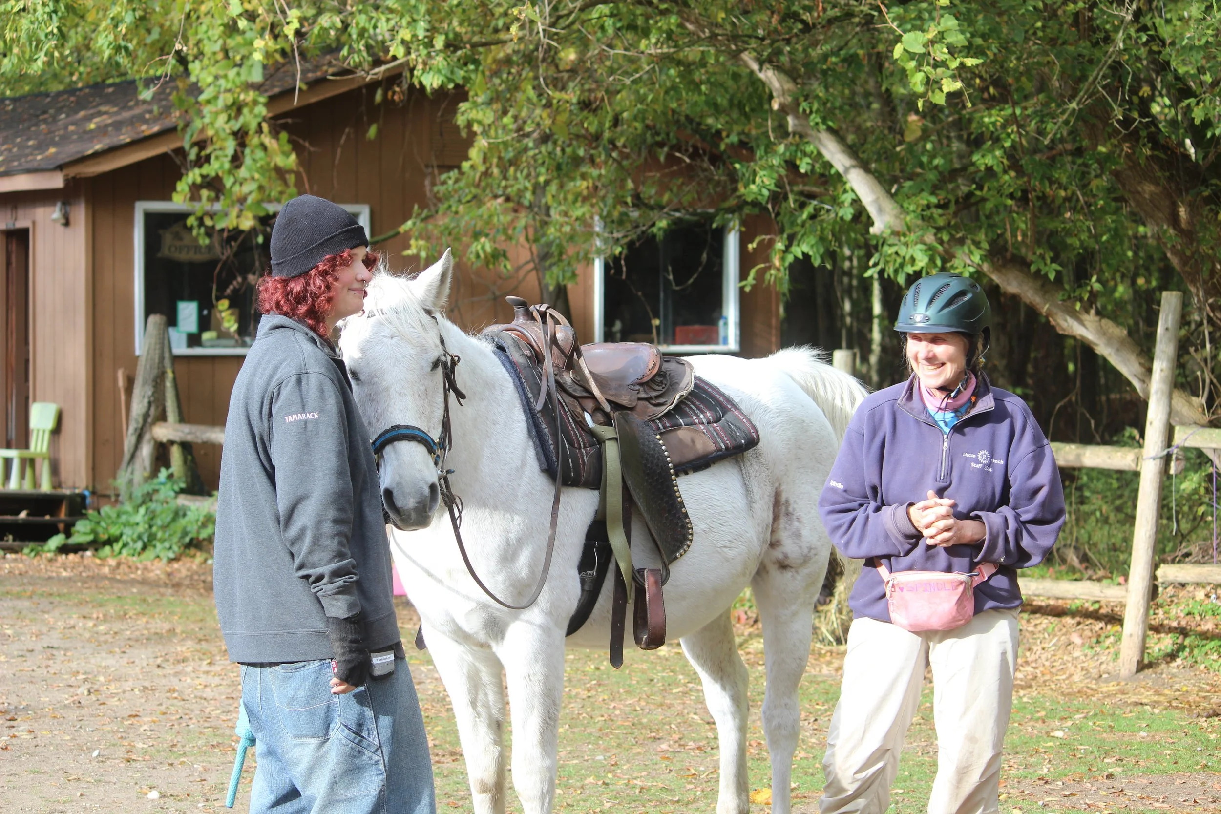 Two women with a white horse outdoors, one woman with reddish hair wearing a black hat and gray jacket, and the other woman with a black riding helmet and navy jacket, smiling at each other. A brown wooden building is in the background.