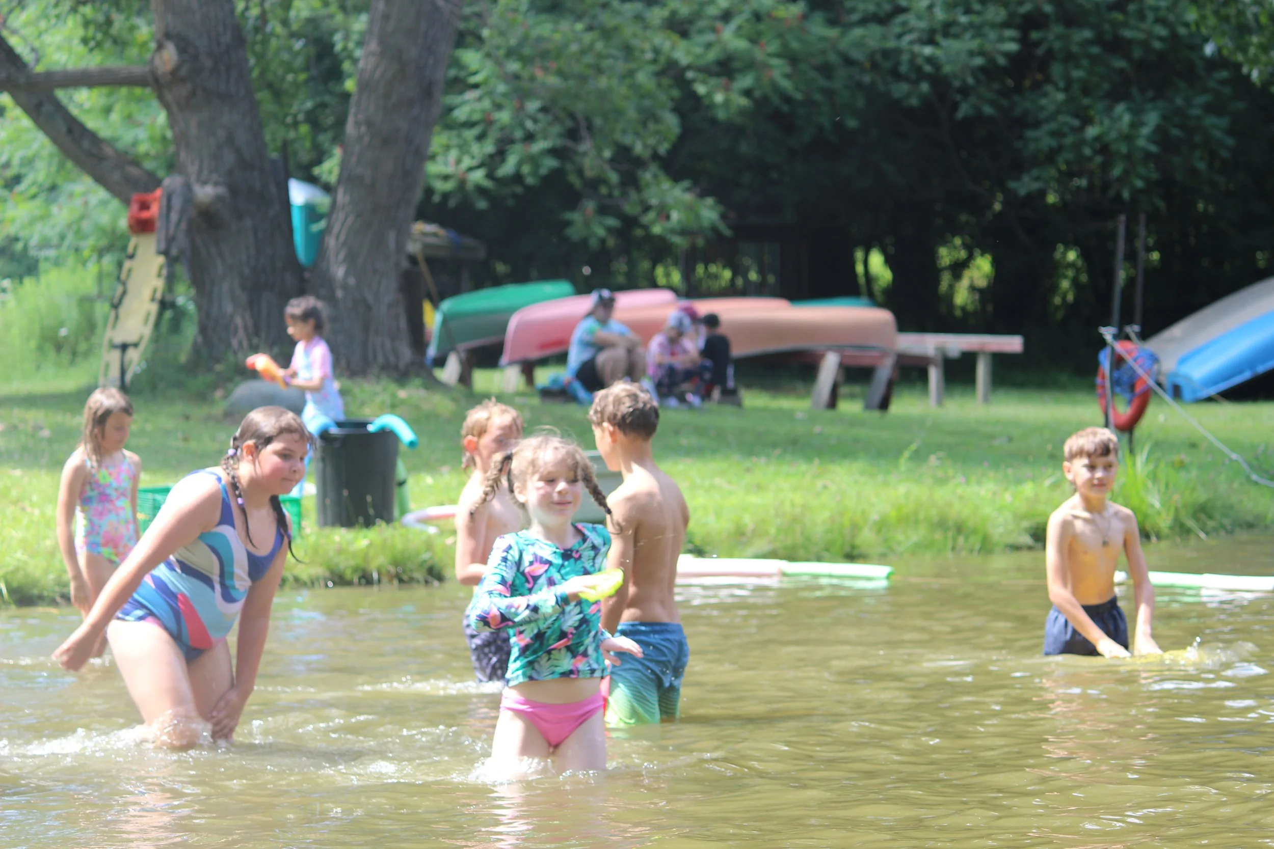 Children playing in a shallow river or pond near a grassy area with trees, some children are wading or standing in the water, and others are near the shore with boats and canoes lined up behind.