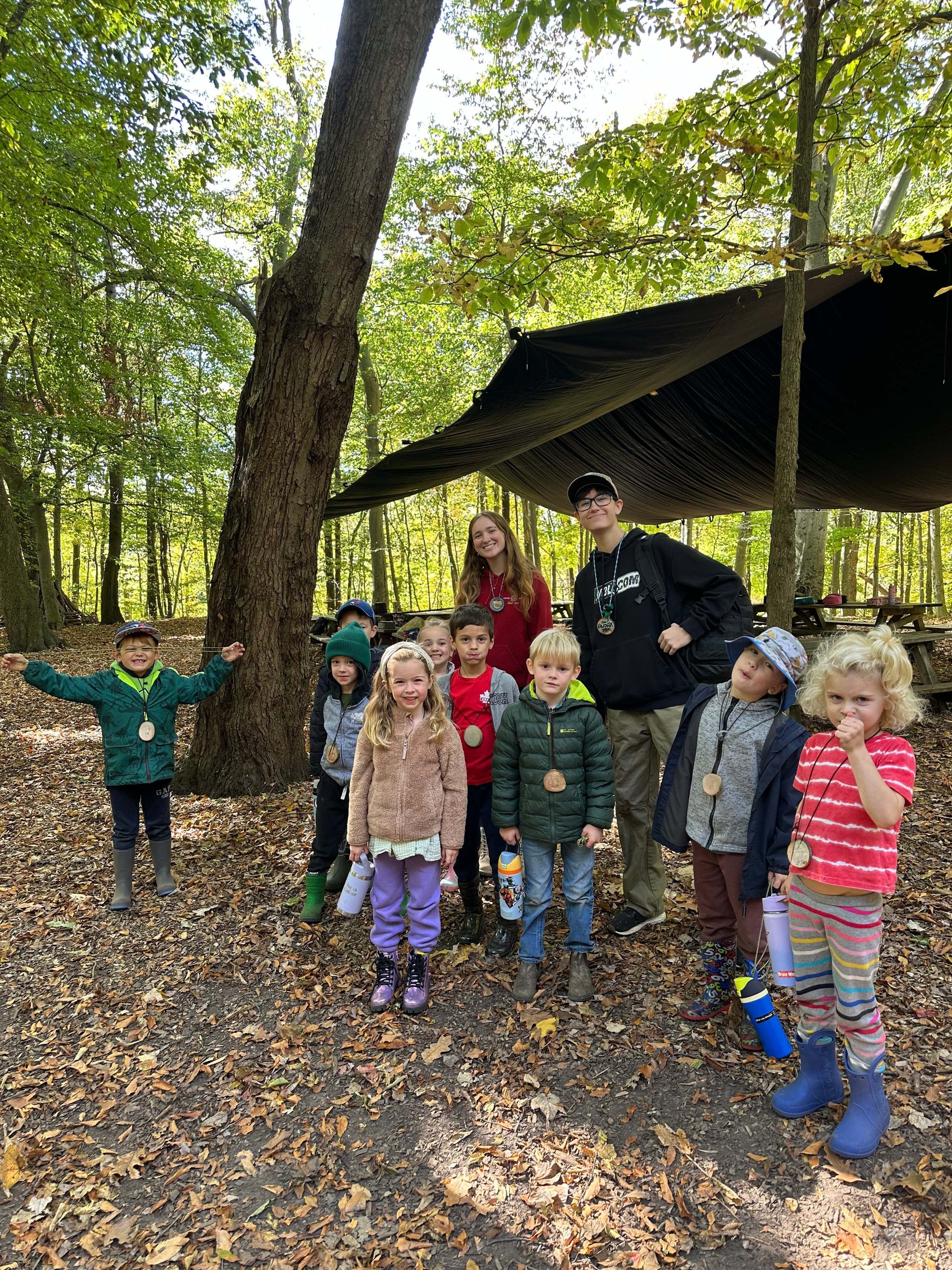Group of children and two adults posing in a wooded outdoor area with leaves on the ground and trees overhead, under a black tarp canopy, some children wearing colorful jackets and boots, holding water bottles, and smiling.