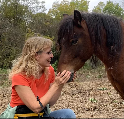A woman with blonde hair and glasses smiling and petting a brown horse outdoors in a natural setting with trees.