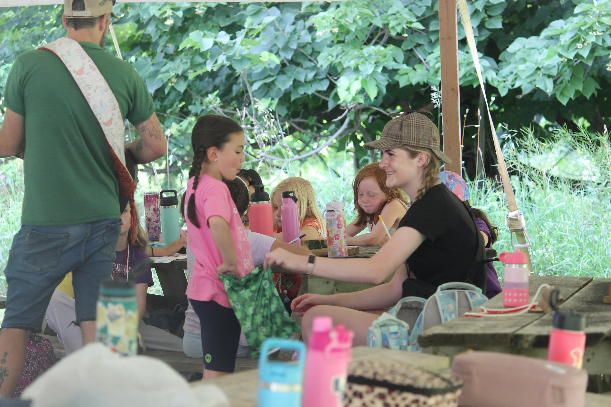 A group of children and a young woman with braided hair, sitting at a wooden picnic table outdoors, surrounded by trees and greenery, with various water bottles and lunch items on the table. The young woman is smiling and wearing a hat, while the chi