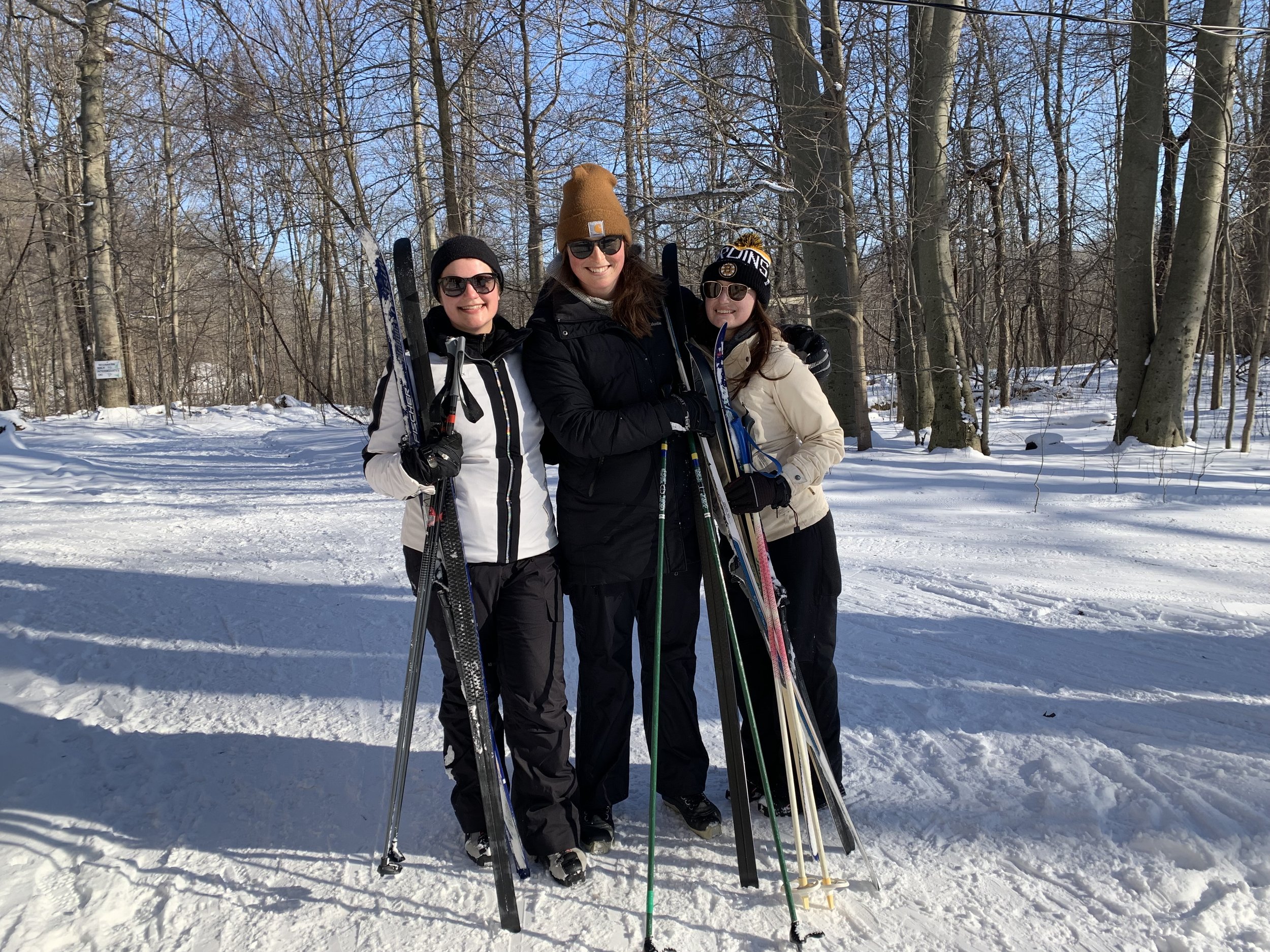Three women in winter clothing standing on snow with skis and ski poles in a wooded area on a sunny day.