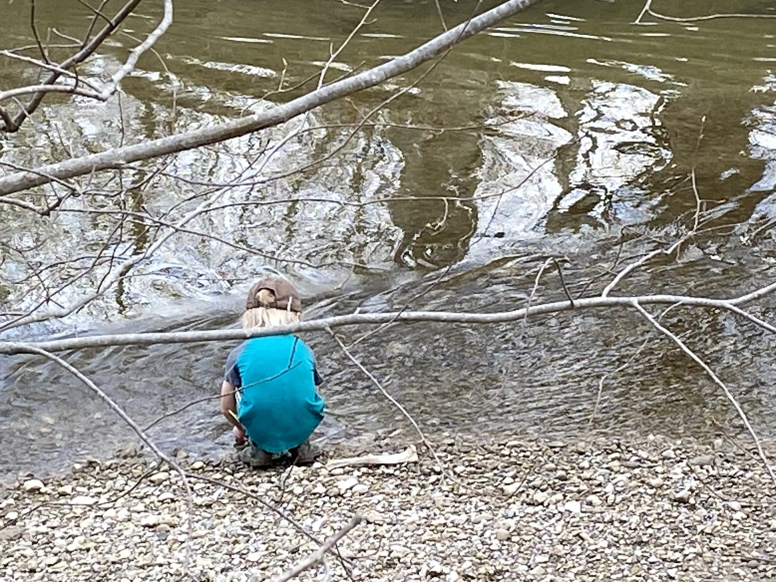 A person crouching at the edge of a stream or river, surrounded by branches and twigs, with water flowing over rocks.