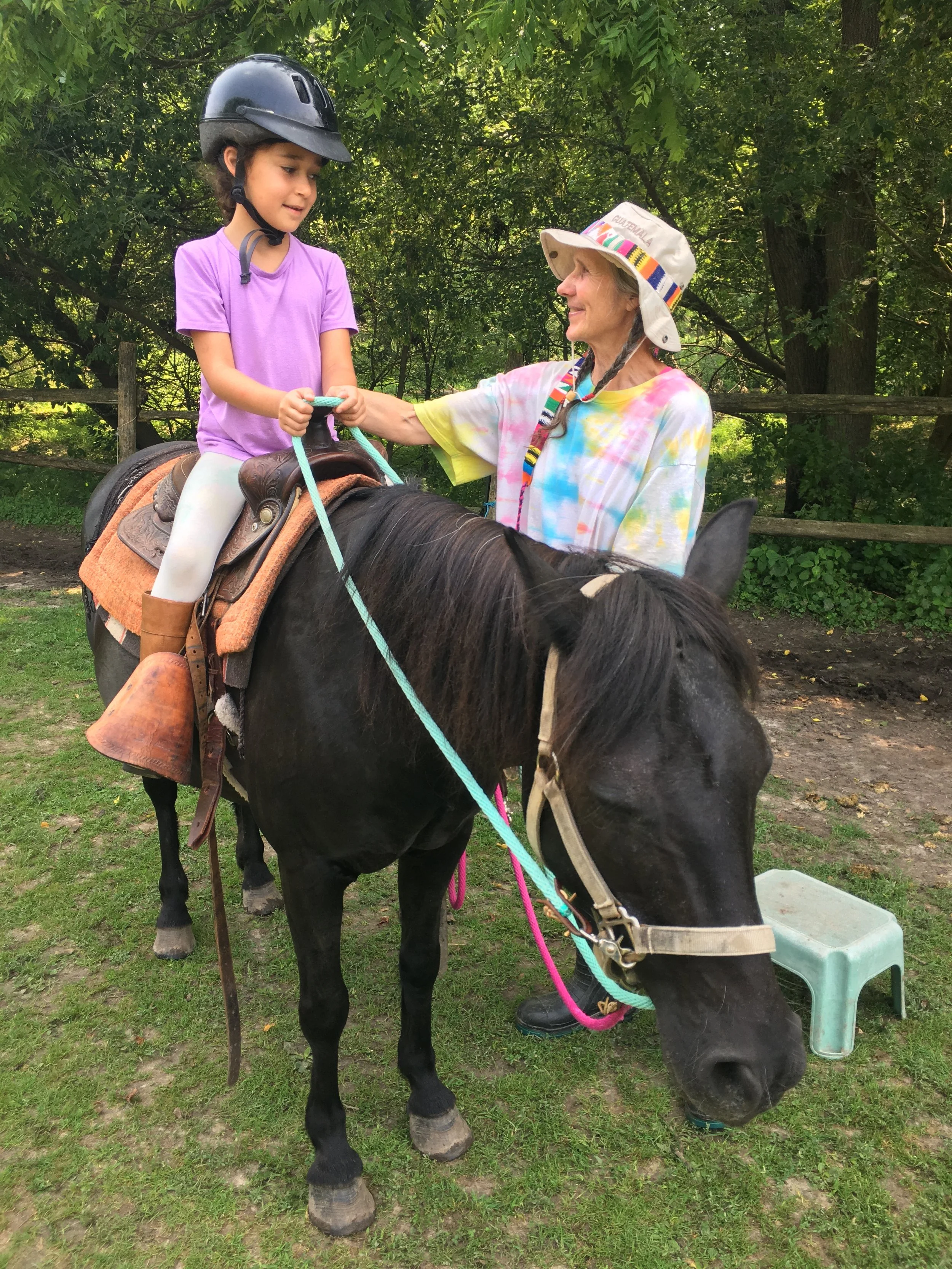 A young girl sitting on a black horse, wearing a grey riding helmet and purple shirt, receiving assistance from an older woman wearing a tie-dye shirt and a colorful hat, outdoors with trees in the background.