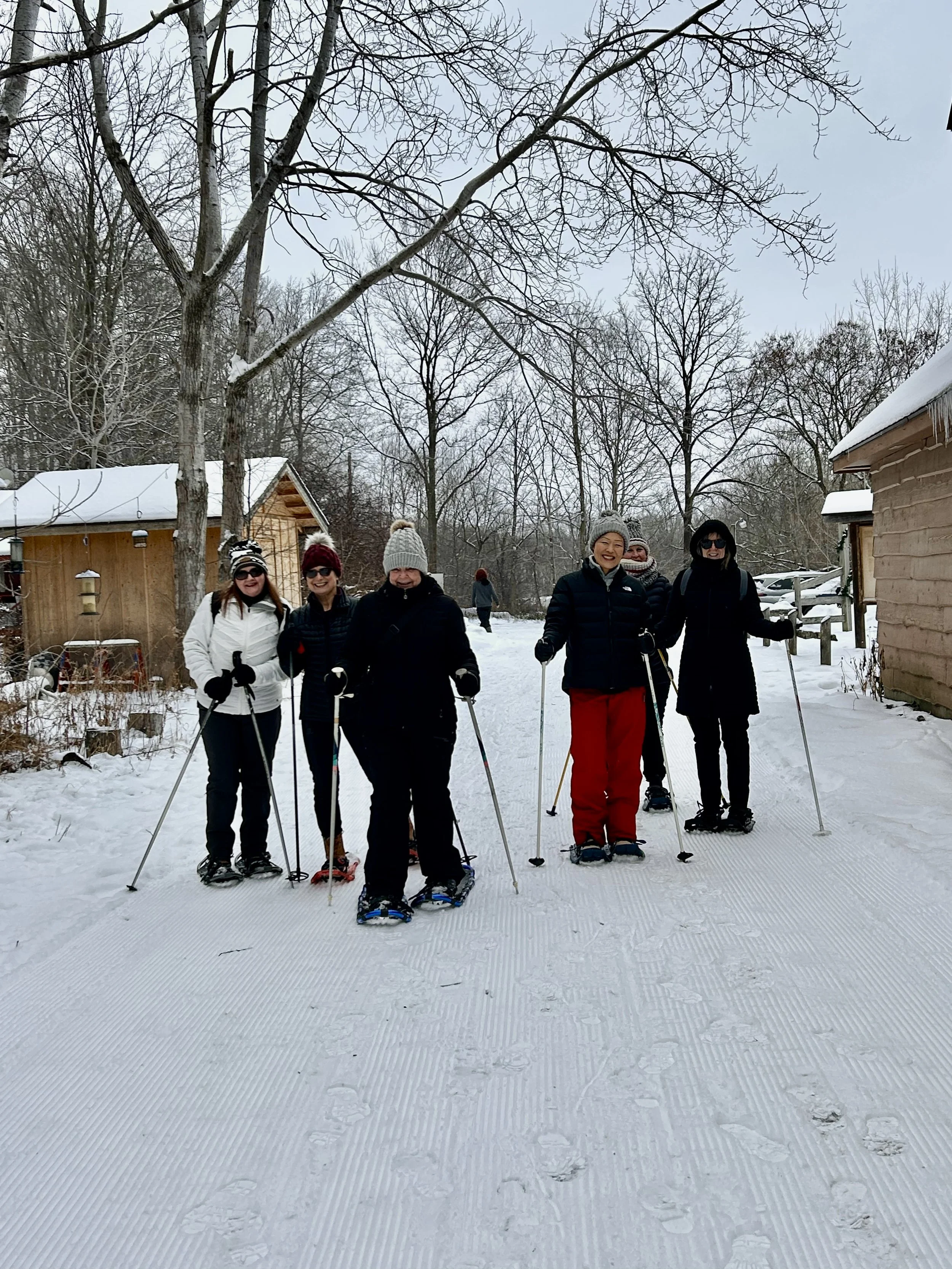 Five women in winter clothing snowshoeing on a snowy path, with trees and small wooden buildings in the background.