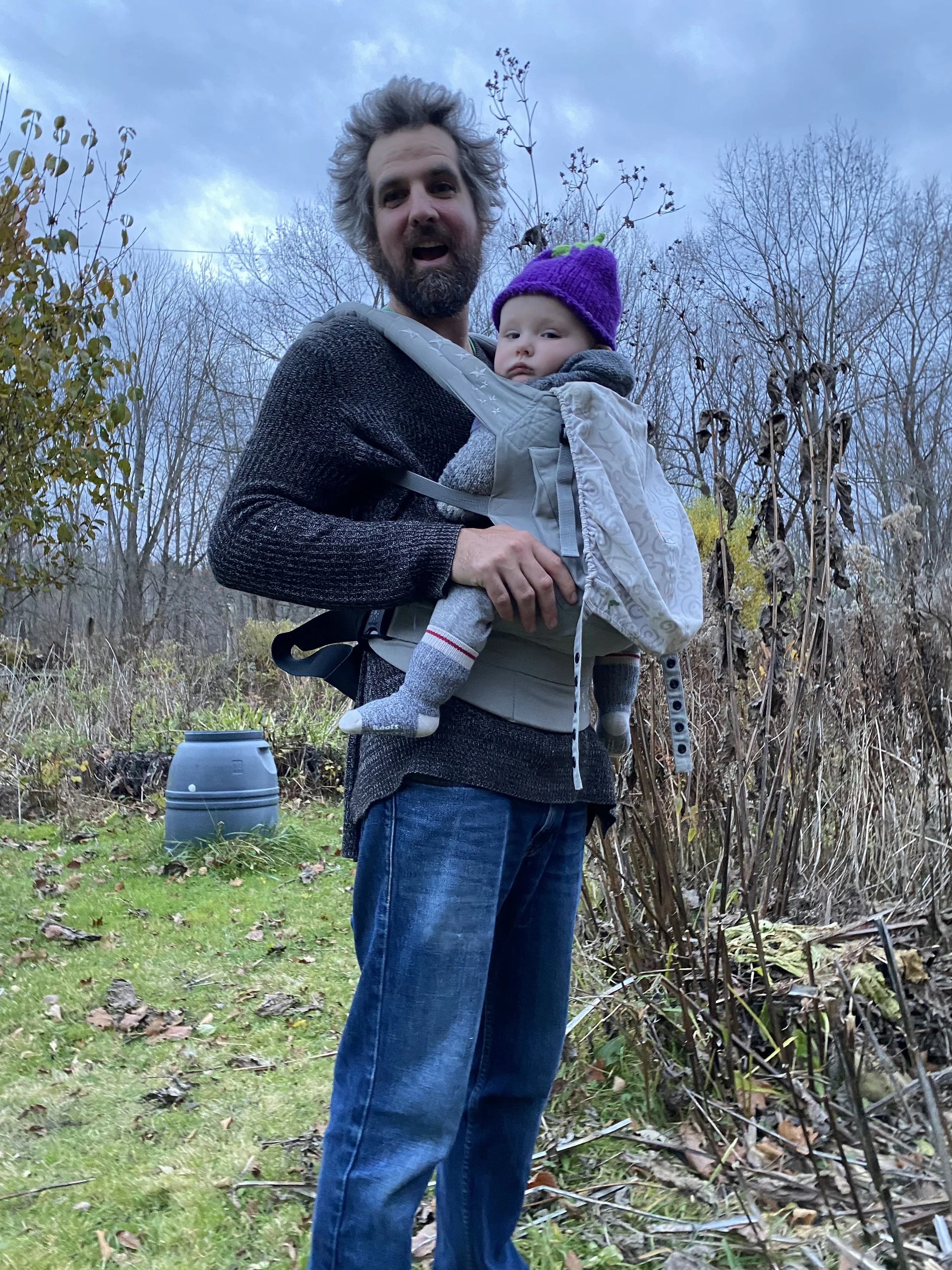 A man with messy hair and beard holding a baby in a gray baby carrier outdoors during late fall or early winter, with bare trees and cloudy sky in the background
