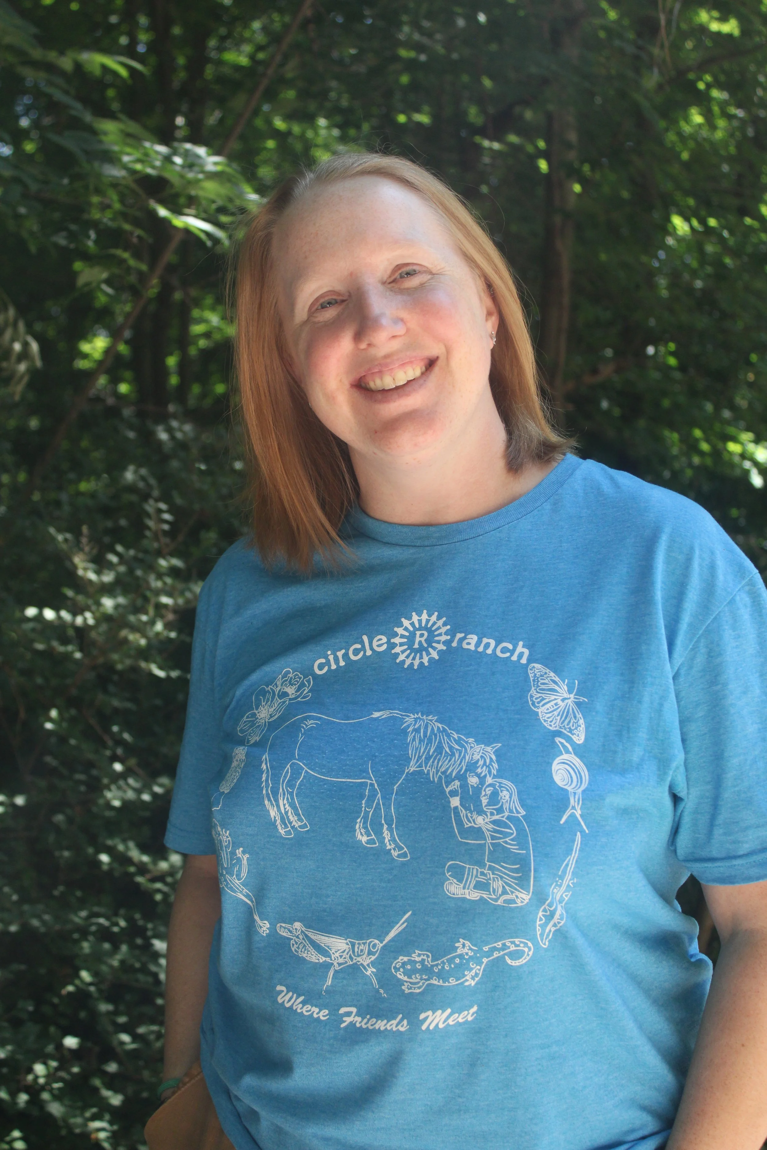A smiling woman with shoulder-length red hair, wearing a blue T-shirt with a wildlife illustration, standing outdoors in a wooded area.