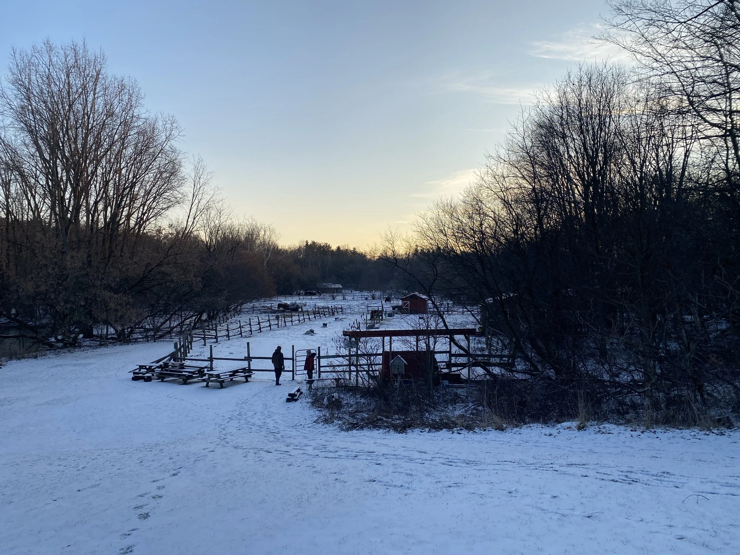 Snow-covered landscape with leafless trees, wooden fences, and a few people walking near a small red barn in the background.