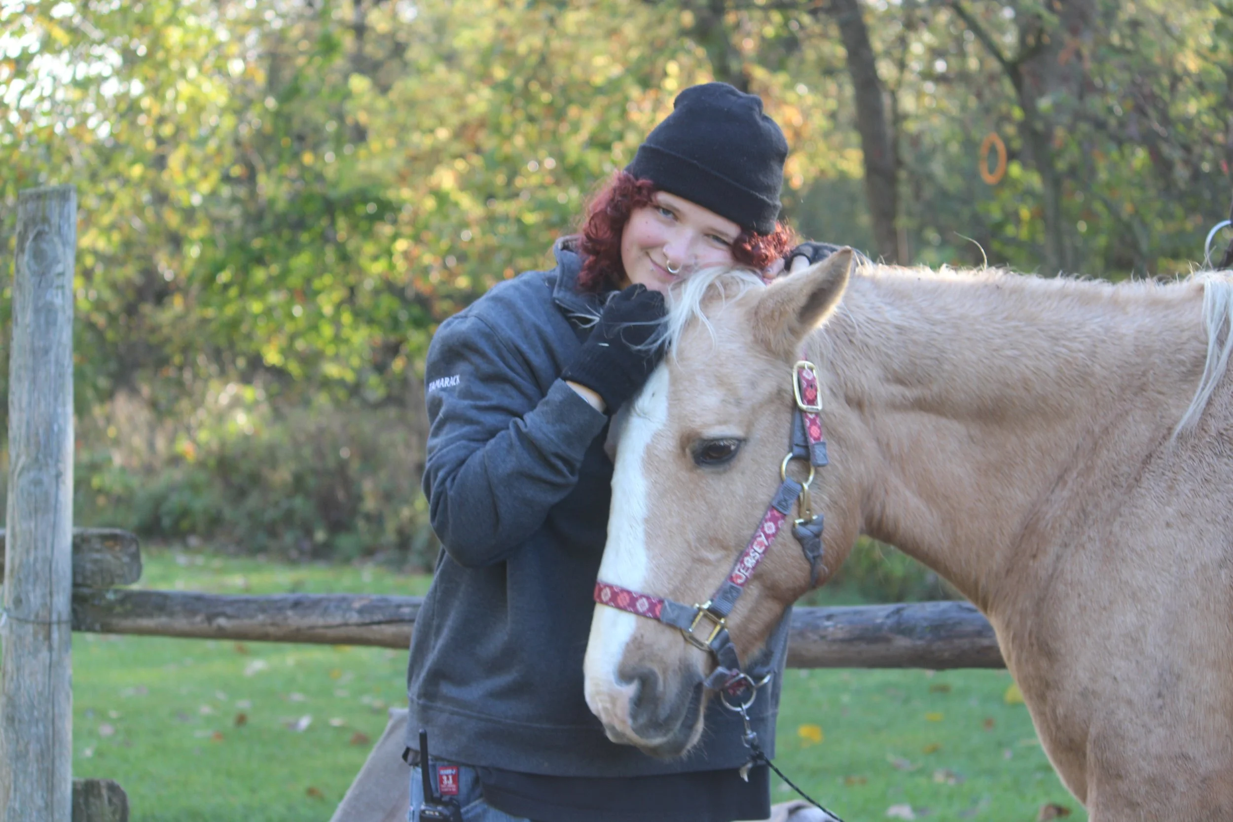 A person with curly red hair, wearing a black beanie, gray hoodie, and black gloves, hugs a light-colored horse with a pink and gray halter in a grassy outdoor setting with trees in the background.