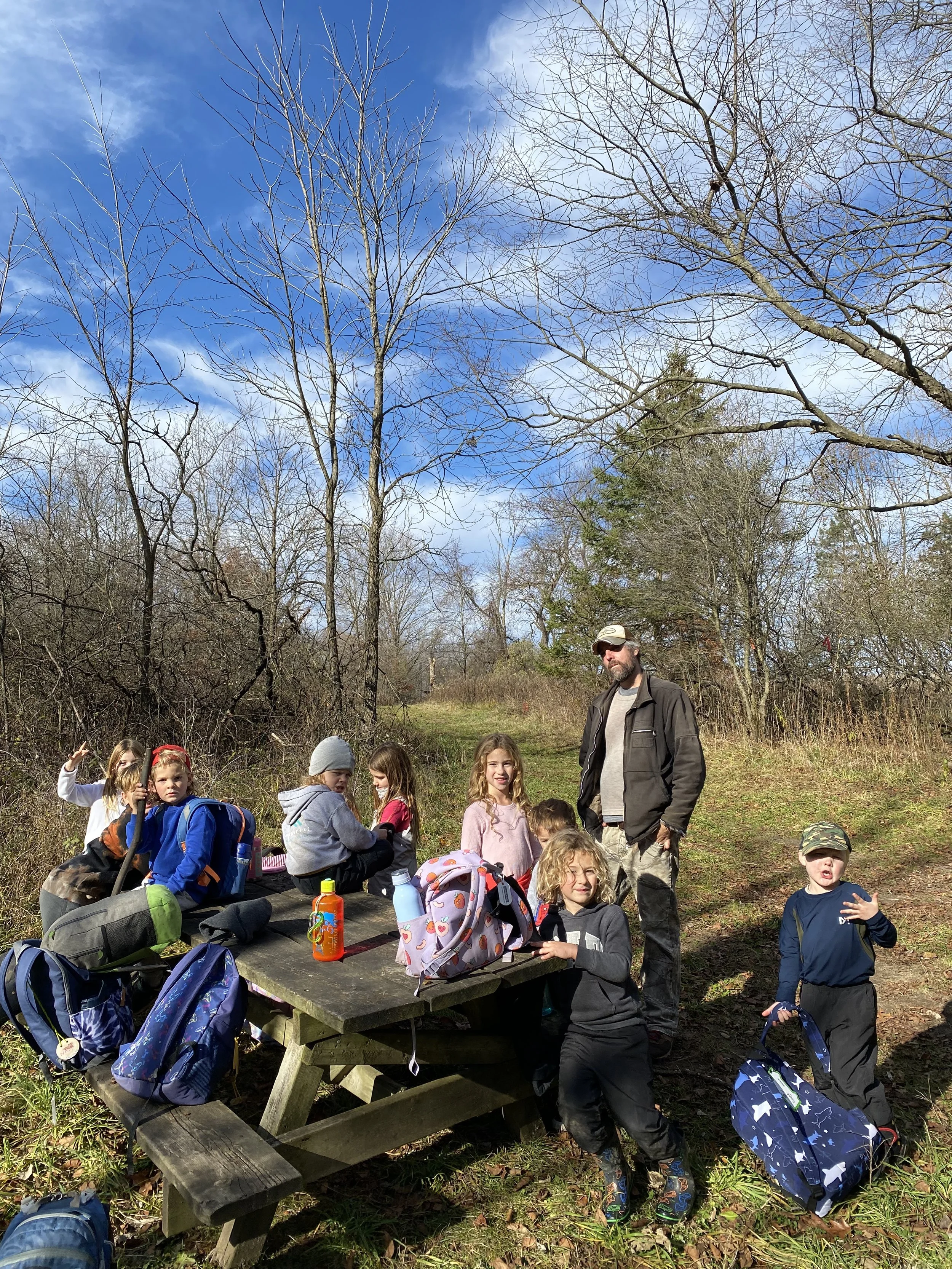 A group of children with a male adult on a hiking trail in a forested area with leafless trees under a partly cloudy blue sky.