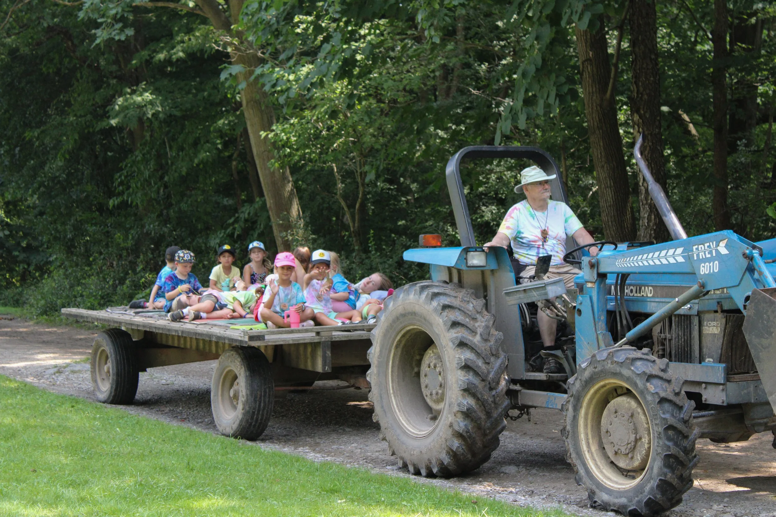 A group of young children sitting on a flatbed trailer being pulled by a blue tractor through a wooded area, supervised by an adult wearing a hat.