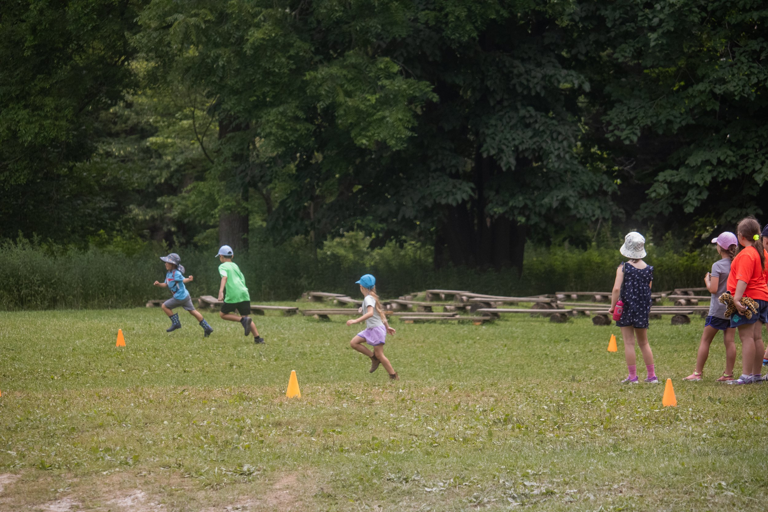 Children running and standing outdoors, some wearing hats, across a grassy field with orange cones set up for an activity, with trees in the background.