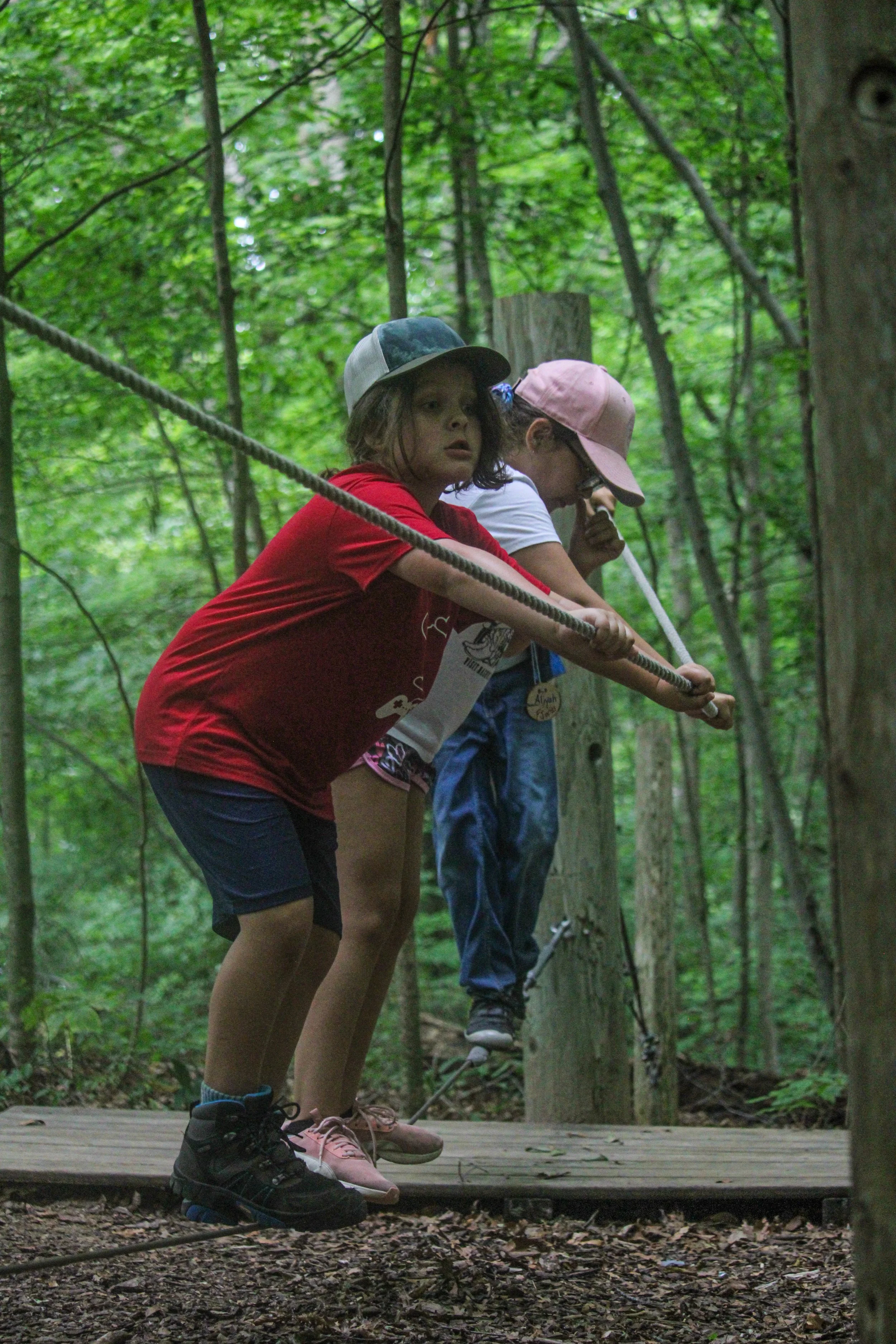 Three children wearing hats and casual clothes walking on a narrow wooden suspension bridge high above the ground in a dense green forest.