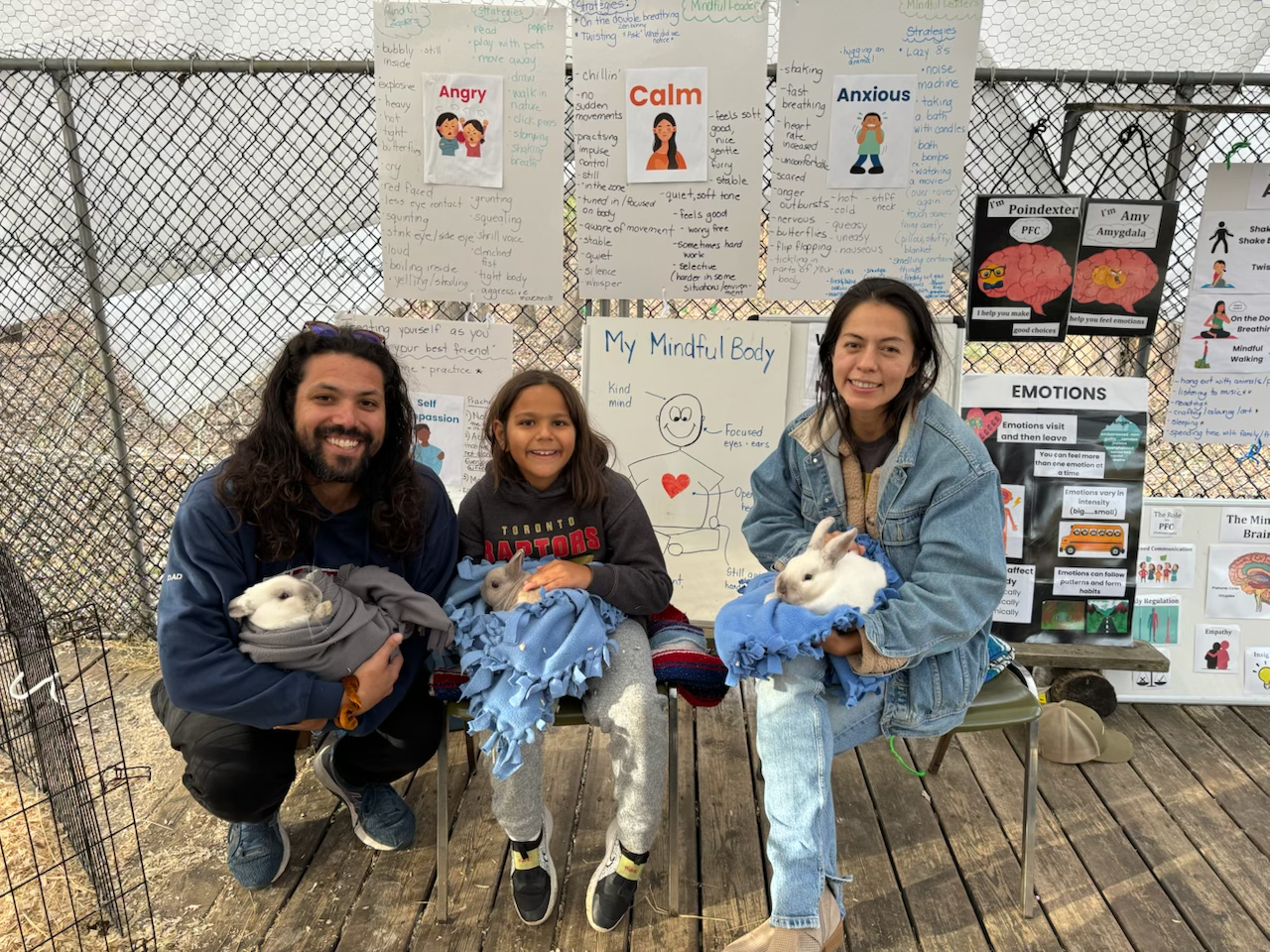 A man and two young girls sitting on chairs inside a fenced outdoor area, each holding a white rabbit wrapped in blue blankets. Behind them are educational posters about emotions and mindfulness, with happy, calm, and anxious face illustrations.