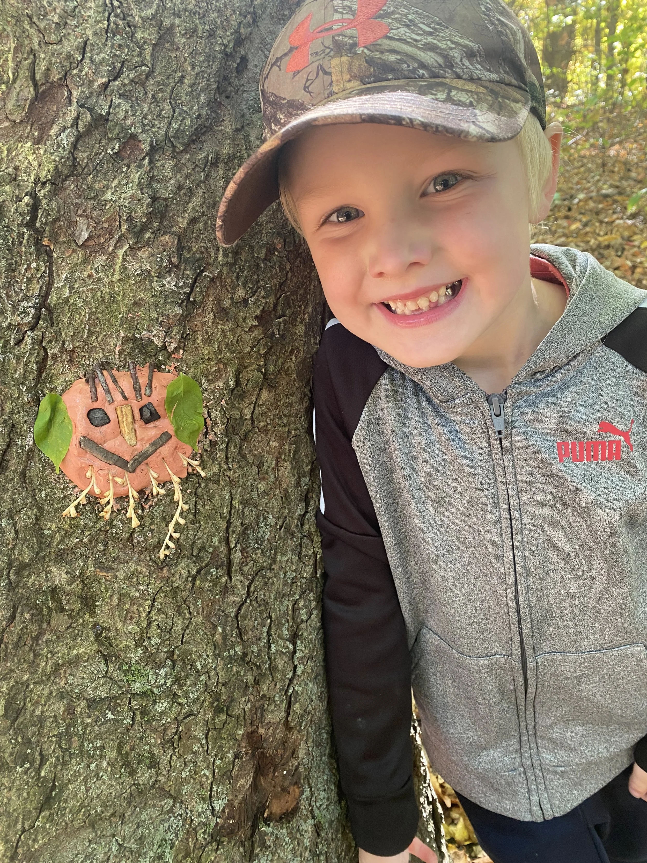 A young boy wearing a camouflage baseball cap and a gray Puma jacket smiles and leans against a tree with an art project on it. The art project, made from a piece of orange fruit or vegetable, has a face drawn with various natural items like leaves, 