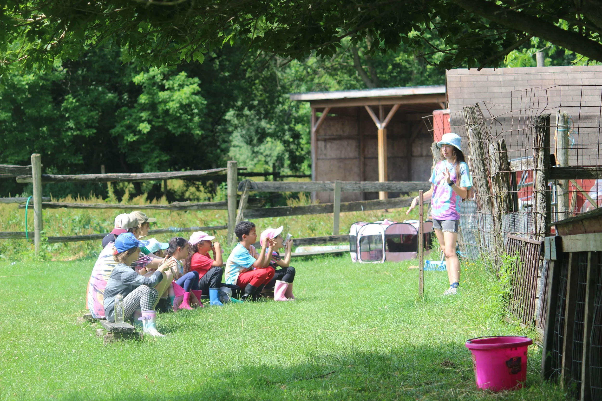 A group of children sitting on a bench outdoors listening to a girl in a tie-dye shirt and helmet speaking.