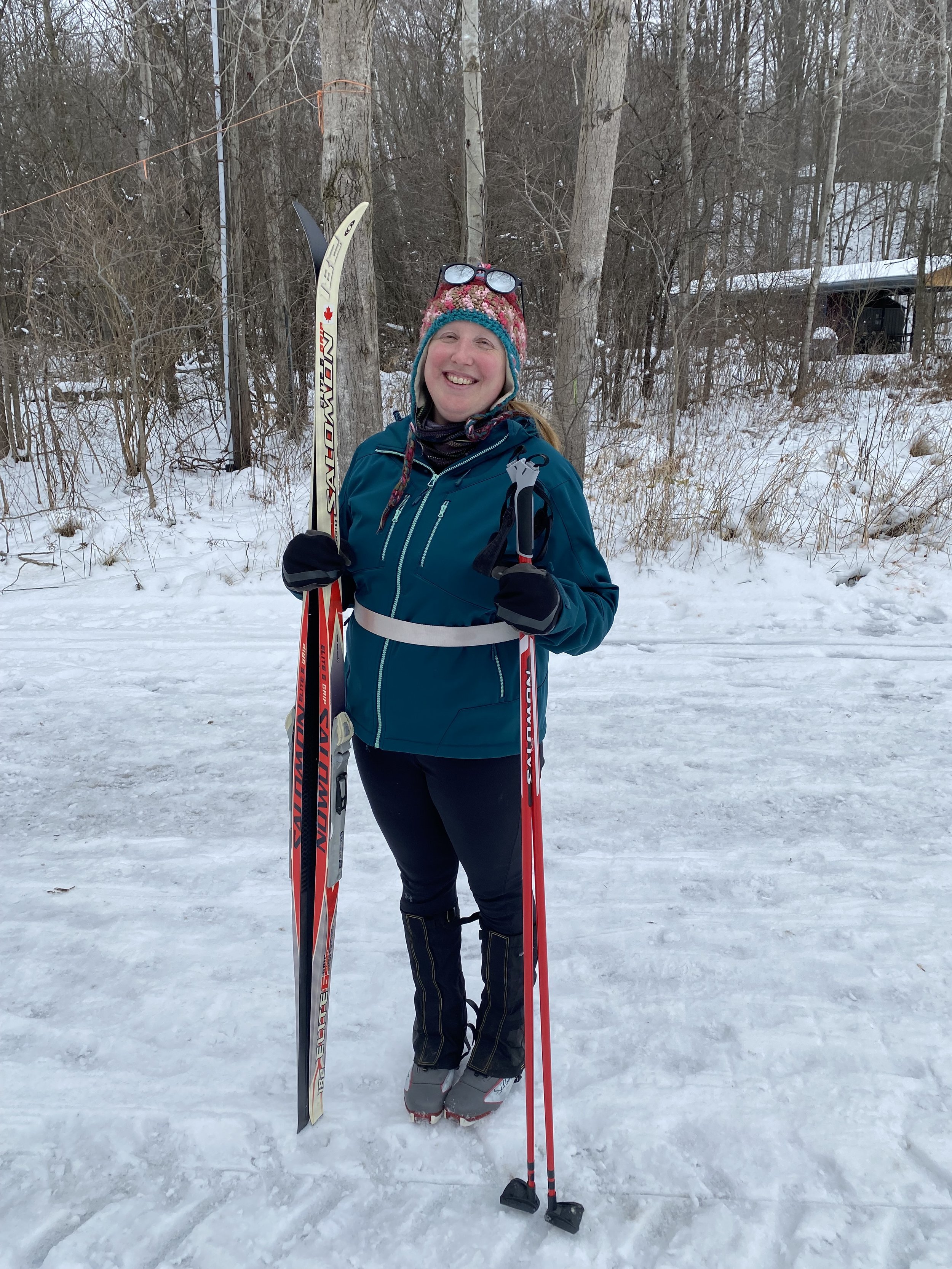 A woman dressed in winter gear holding cross-country skis and poles, standing on snow in a wooded area, smiling.