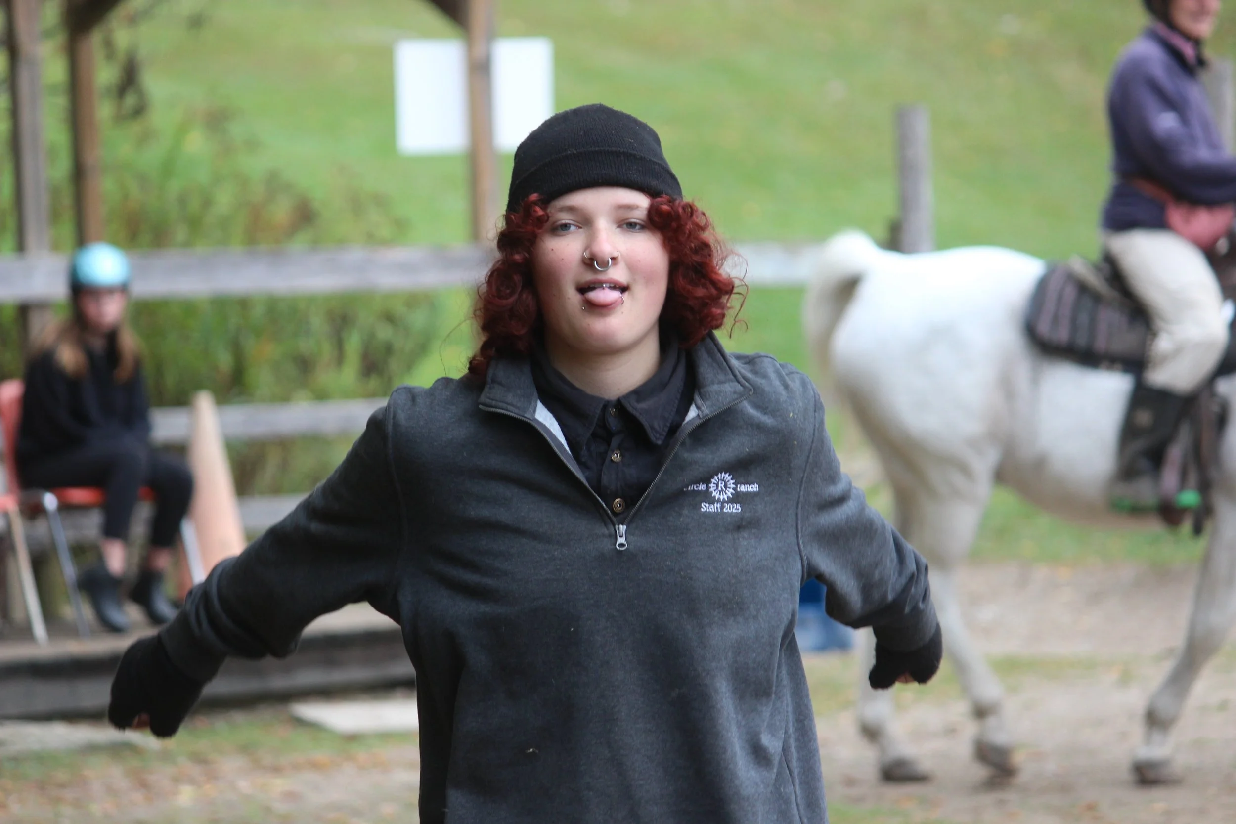 Young woman with red curly hair and septum piercing, wearing a black beanie and gray zip-up jacket, standing outdoors with a playful expression including sticking out her tongue. In the background, there is a woman in a helmet sitting on a chair and a person riding a horse with white fur and saddle.
