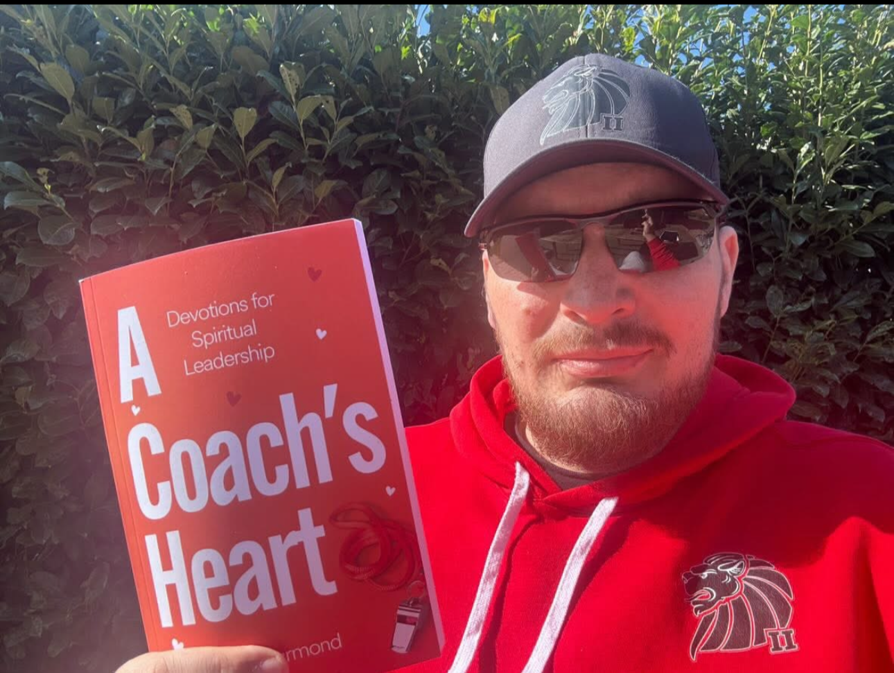 A man wearing sunglasses, a gray cap with a lion logo, and a red hoodie with a matching lion logo holds a book titled 'A Coach's Heart: Devotions for Spiritual Leadership' in front of a green bush.
