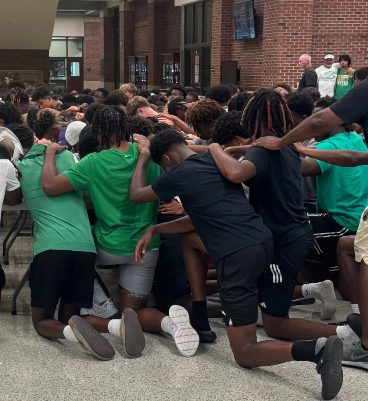 A large group of young people praying together, kneeling and with hands on each other's shoulders in a prayer circle indoors, with some adults standing in the background.