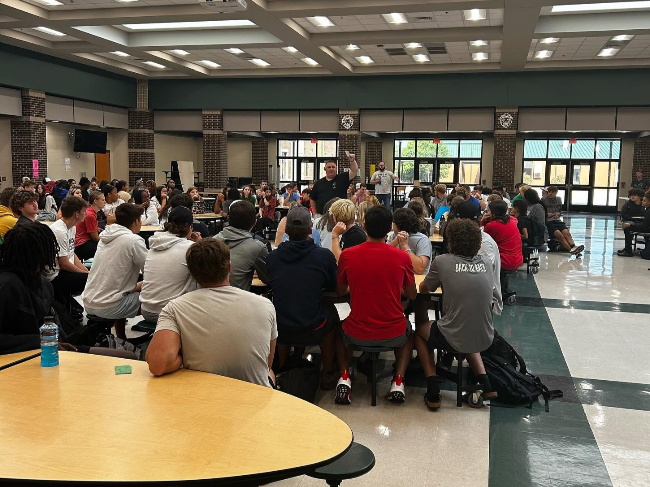 A large group of students seated at tables in a school cafeteria listening to a speaker who is standing and gesturing. The room has high ceilings, large windows, and brick columns.