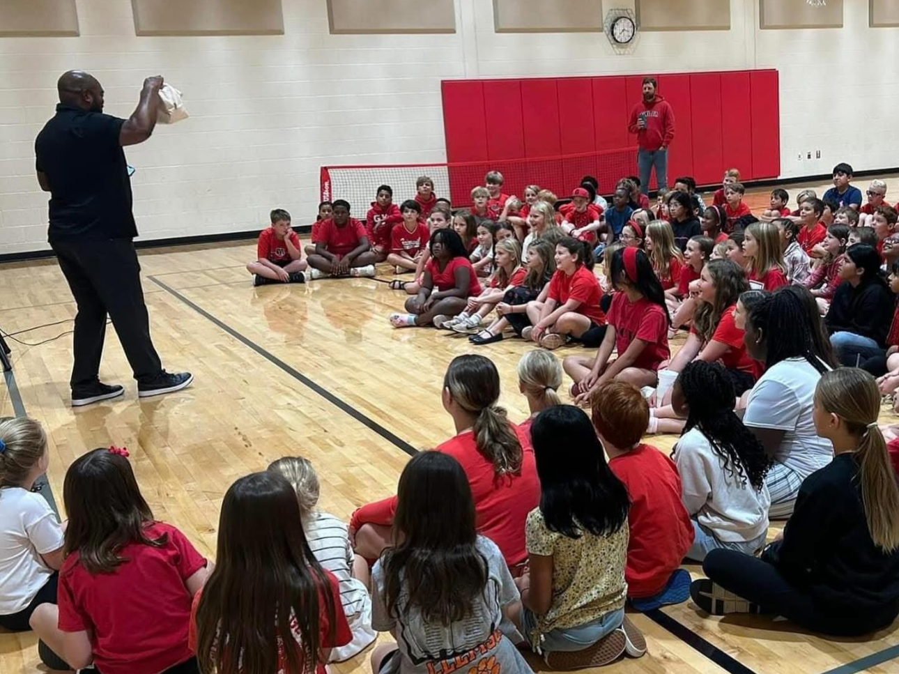 A man giving a presentation to a large group of children sitting on the gymnasium floor, with most children wearing red shirts, in a school gymnasium with a hardwood floor, red padding on the wall, a small soccer goal, and a clock on the wall.