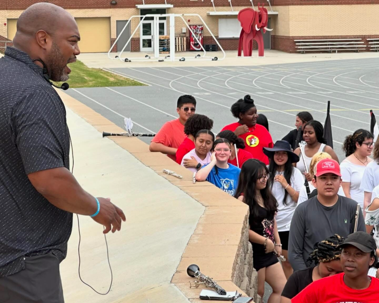 A man speaking to a group of students outdoors on a school track field, with climbing poles and a red elephant sculpture in the background.