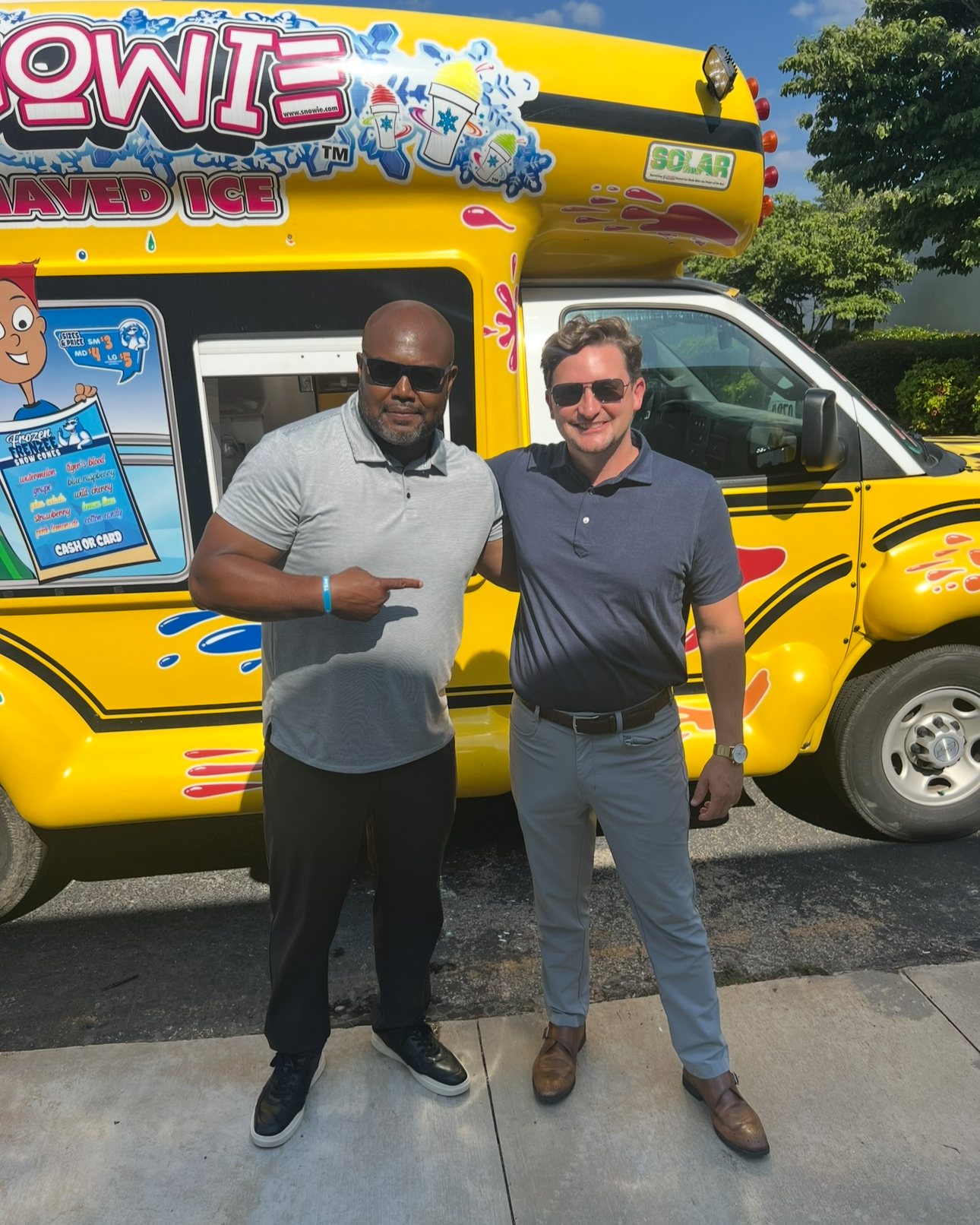 Two men smiling and posing in front of a yellow snow cone truck with colorful decals. One man points at the other.