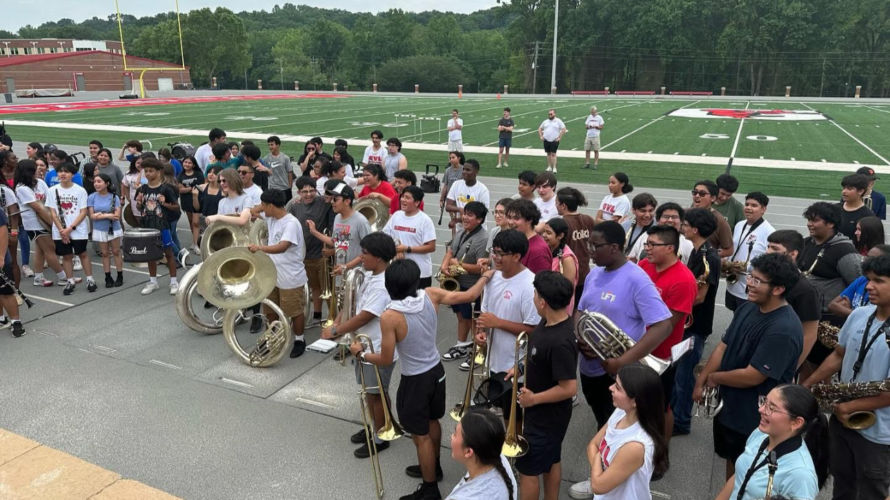 A large group of students and musicians gathered on an outdoor basketball court near a football field, with some holding musical instruments like tubas, saxophones, and trumpets, watching or preparing for a band performance.