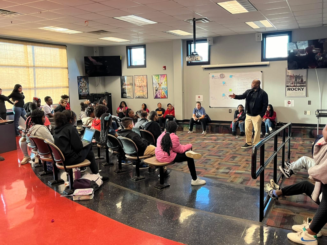A classroom with students seated and a teacher standing at the front, engaging with the class. The room has several posters on the walls, large windows, and a whiteboard.