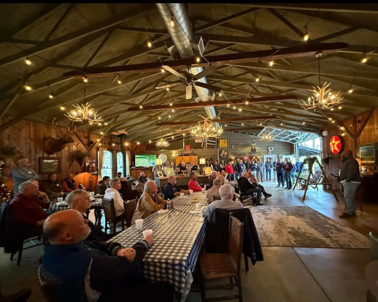 Indoor gathering in a rustic wooden hall with people seated at tables, a speaker on stage, and diverse decorations including antler chandeliers and mounted elk head, illuminated by string lights and chandeliers.