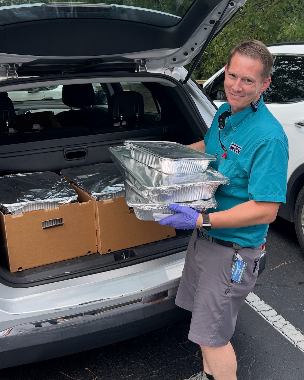 A man in a blue uniform and purple gloves loading foil-covered food containers into the back of a vehicle during daytime.