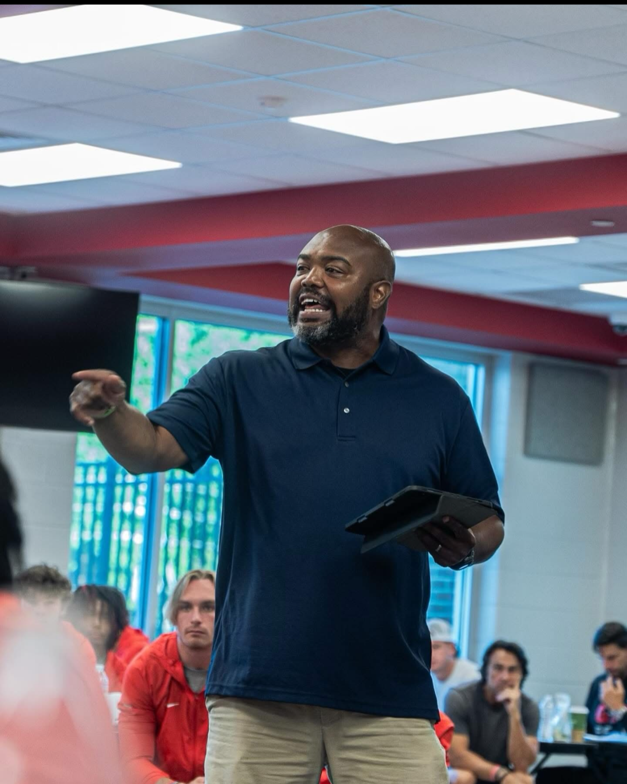 A man speaking passionately in a classroom or meeting room with students seated at tables, some wearing red jackets, and one person with blond hair visible in the background.