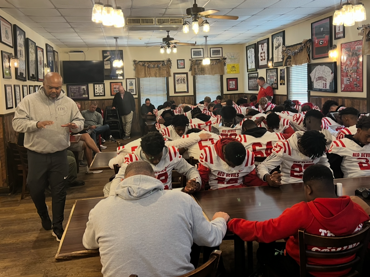 Football team in uniform praying together in a restaurant with framed sports memorabilia on the walls and overhead lighting.